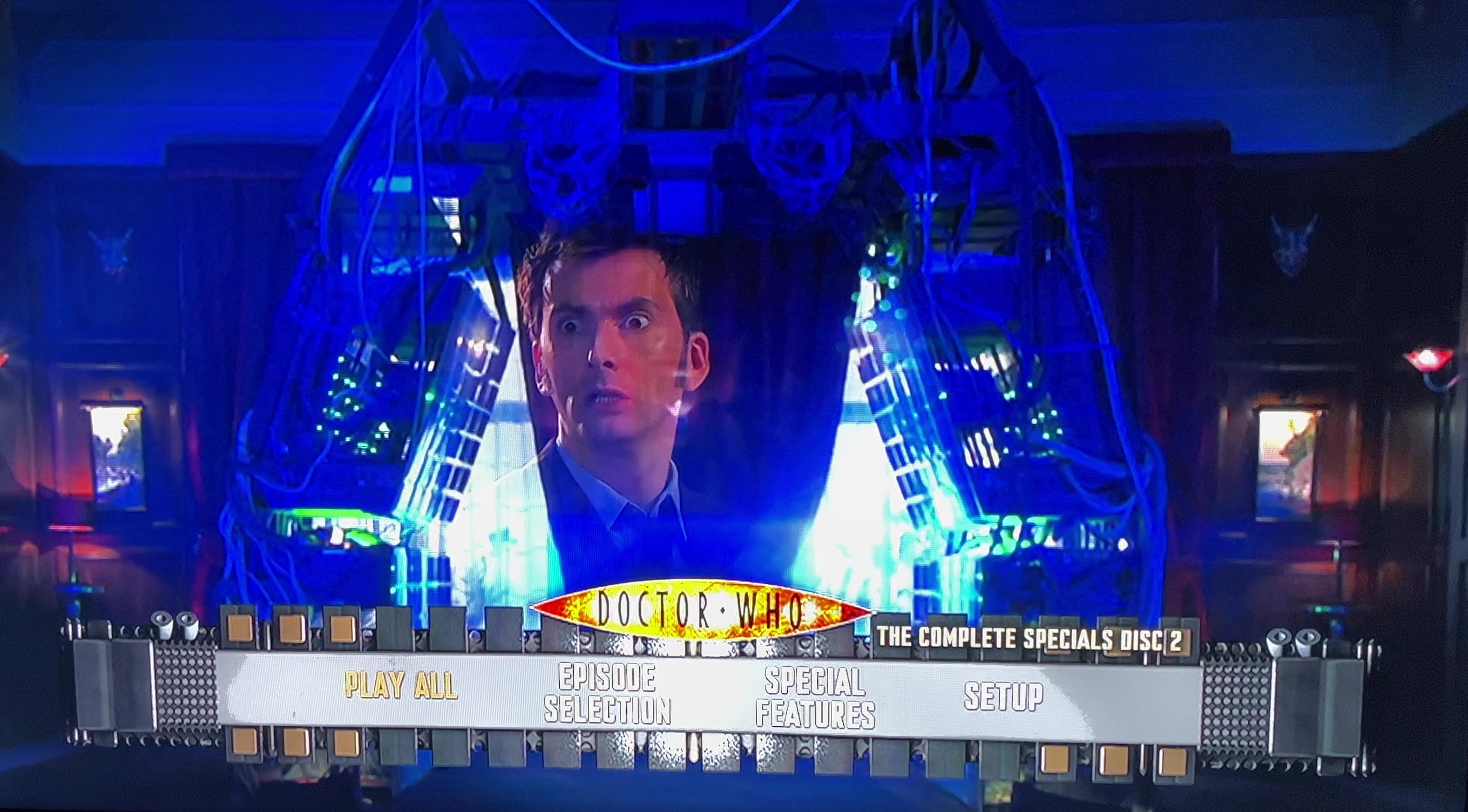 The menu for Disc 2 of the Doctor Who 2008 to 2010 specials Blu-ray steelbook. The screen shows David Tennant as the 10th Doctor looking concerned, his image shown within the Immortality Gate machine used in the series finale. The menu options are across the bottom of the screen, with the Doctor Who logo just above them.