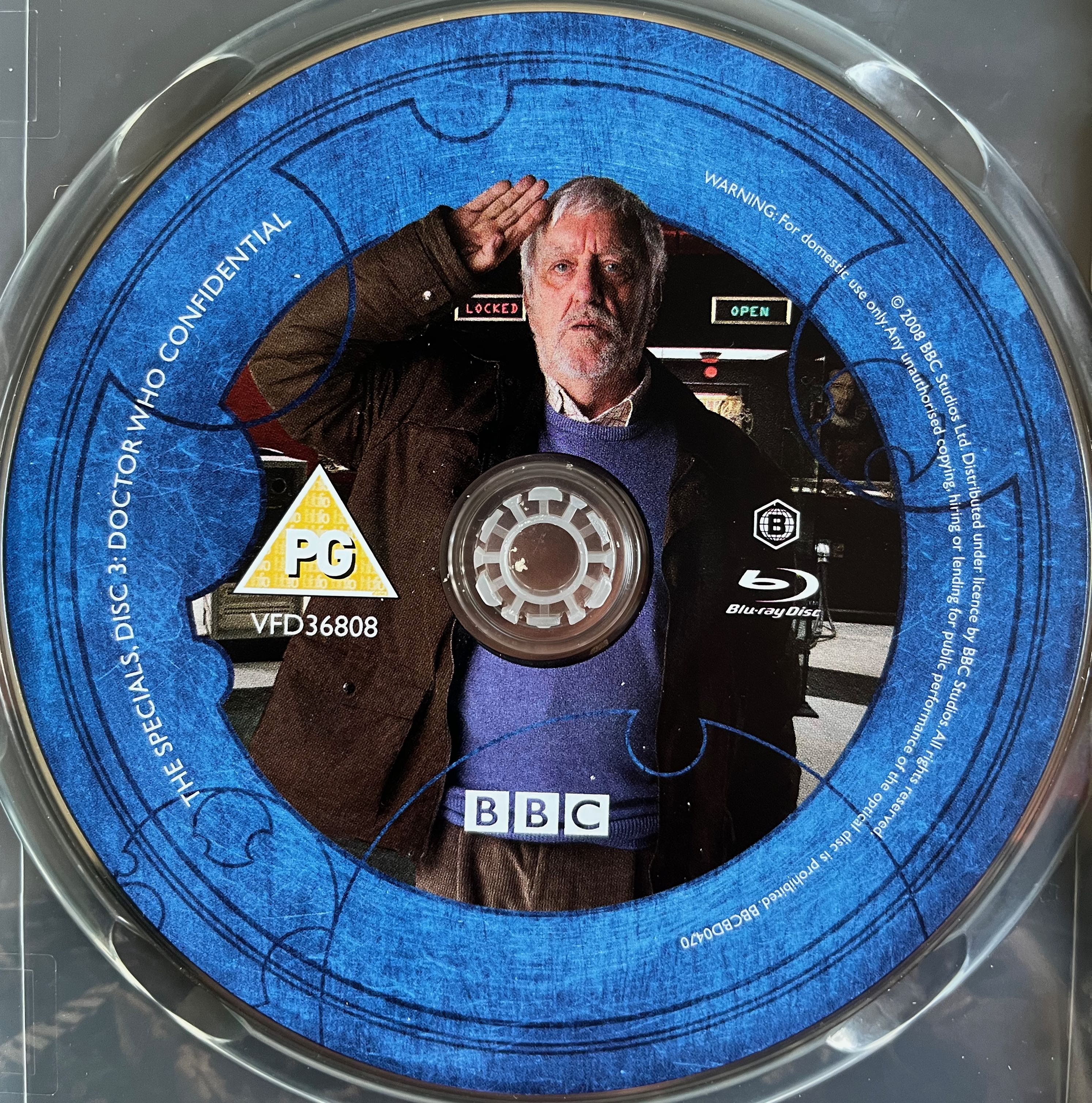 Disc 3 in the Doctor Who 2008 to 2010 Specials Blu-ray set. It features a photo of Bernard Cribbins as Wilfred standing to attention and saluting, while behind him 2 cabinets have signs saying locked and open respectively. A blue border around the edge of the disc states that it contains Doctor Who Confidential and provides copyright information.
