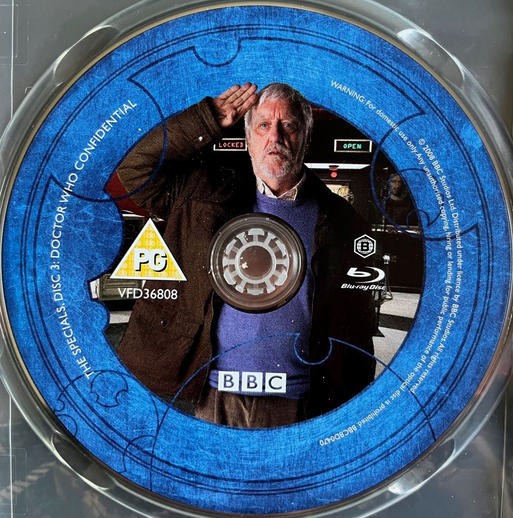 Disc 3 in the Doctor Who 2008 to 2010 Specials Blu-ray set. It features a photo of Bernard Cribbins as Wilfred standing to attention and saluting, while behind him 2 cabinets have signs saying locked and open respectively. A blue border around the edge of the disc states that it contains Doctor Who Confidential and provides copyright information.