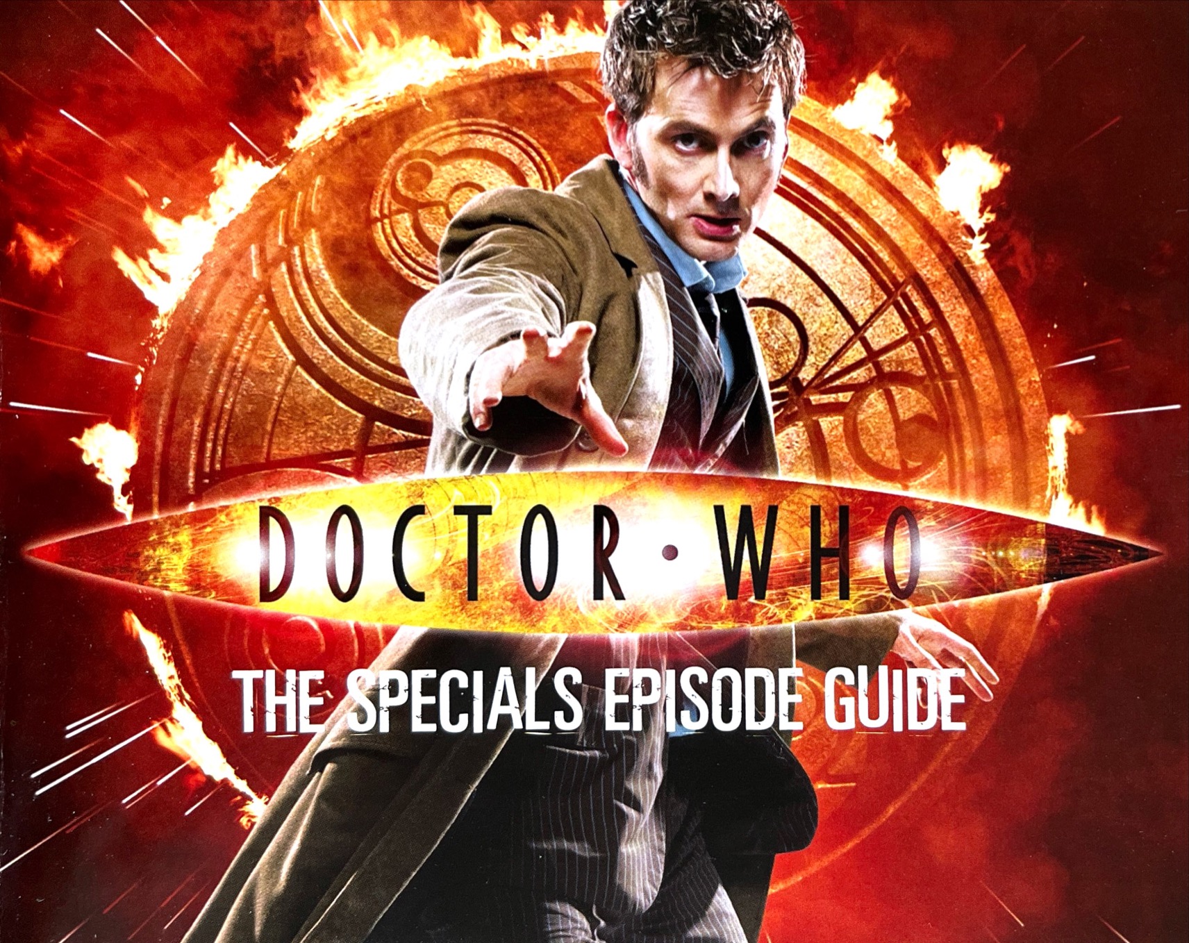 The cover of the Doctor Who 2008-2010 Specials Episode Guide booklet included with the original DVD release. It shows a photo of David Tennant's Doctor wearing a flowing brown jacket over a pinstripe suit, and reaching out towards us with his right hand, while behind him is a large golden circle covered in Gallifreyan symbols with flames erupting around the edge. Over the image is the Doctor Who logo and the title of the booklet in white text, The Specials Episode Guide.
