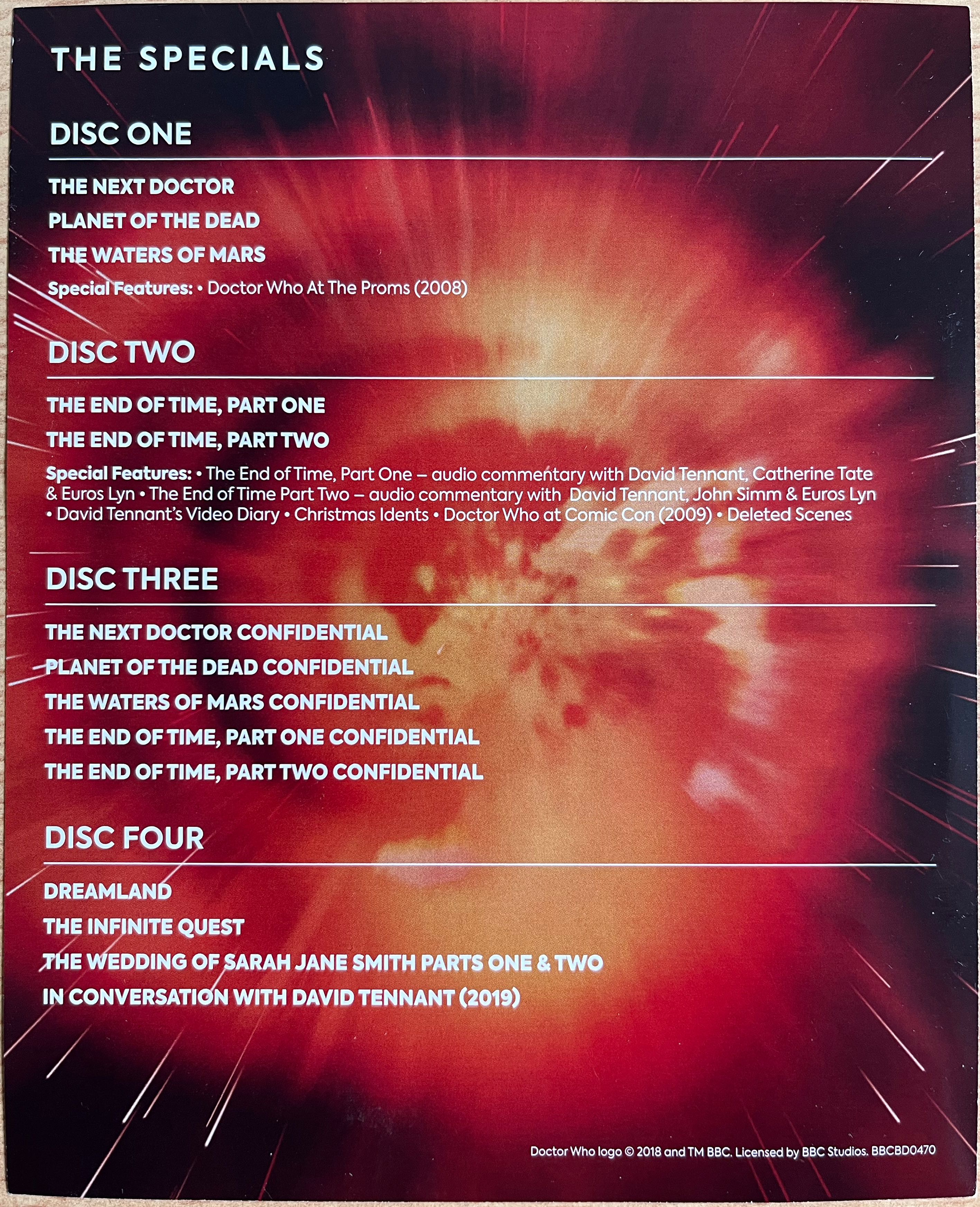 Rear side of the flyer included with the Doctor Who 2008 to 2010 Specials Blu-ray Steelbook, showing the list of episodes and extras on each of the 4 discs, in white text over a red and orange explosion of light that could be an entrance to the time vortex, given the white lights of stars forming lines of movement around it.