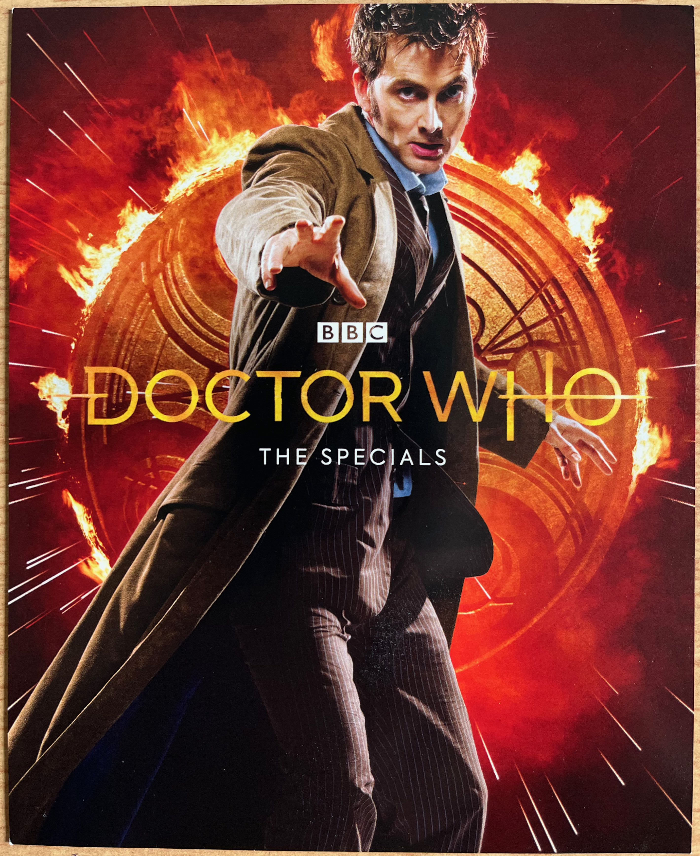 Front side of the flyer included with the Doctor Who 2008 to 2010 Specials Blu-ray Steelbook, featuring a photo of David Tennant's Doctor wearing a flowing brown jacket over a pinstripe suit, and reaching out towards us with his right hand, while behind him is a large golden circle covered in Gallifreyan symbols with flames erupting around the edge.