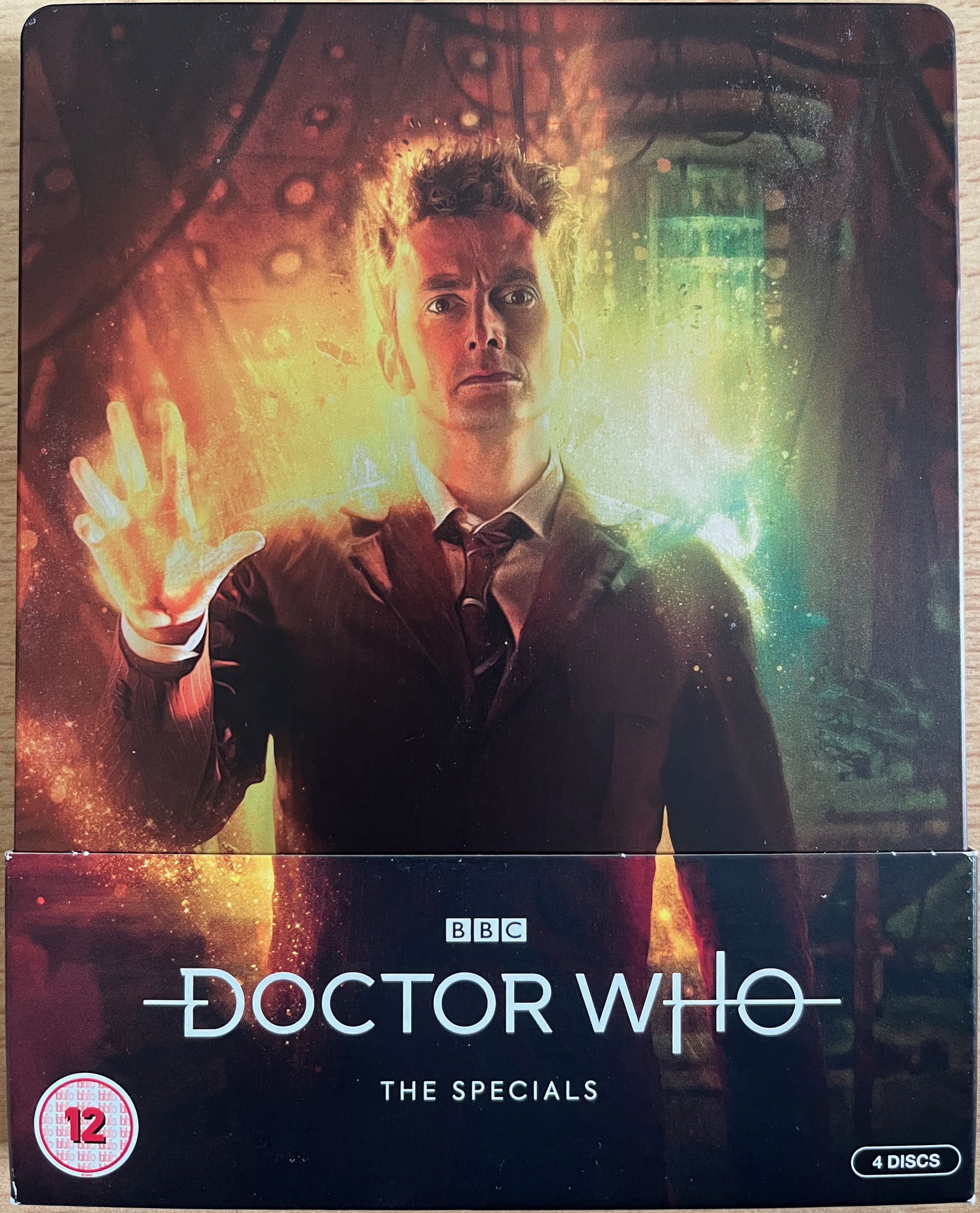 The front cover of the Doctor Who 2008 to 2010 Specials Blu-ray steelbook, which is sitting in a cardboard pocket that has the Doctor Who logo on it. The steelbook cover shows David Tennant as the Doctor in a brown suit, standing in the Tardis, with glowing yellow regeneration energy around him.