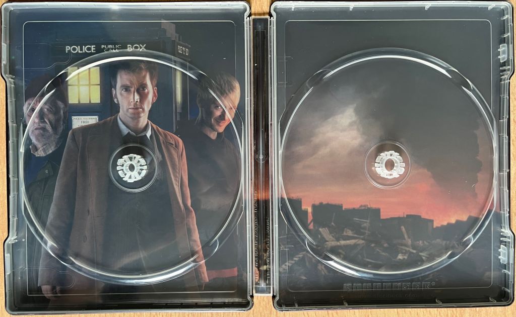The interior spread of the Doctor Who 2008 to 2010 specials Blu-ray steelbook, behind where the discs are stored. The left side shows David Tennant as the Doctor, Bernard Cribbins as Wilfred and John Simm grinning as the Master, standing in front of the Tardis. The right side shows a waste ground, as if buildings have been destroyed, in front of a red horizon and dark grey clouds in the sky.