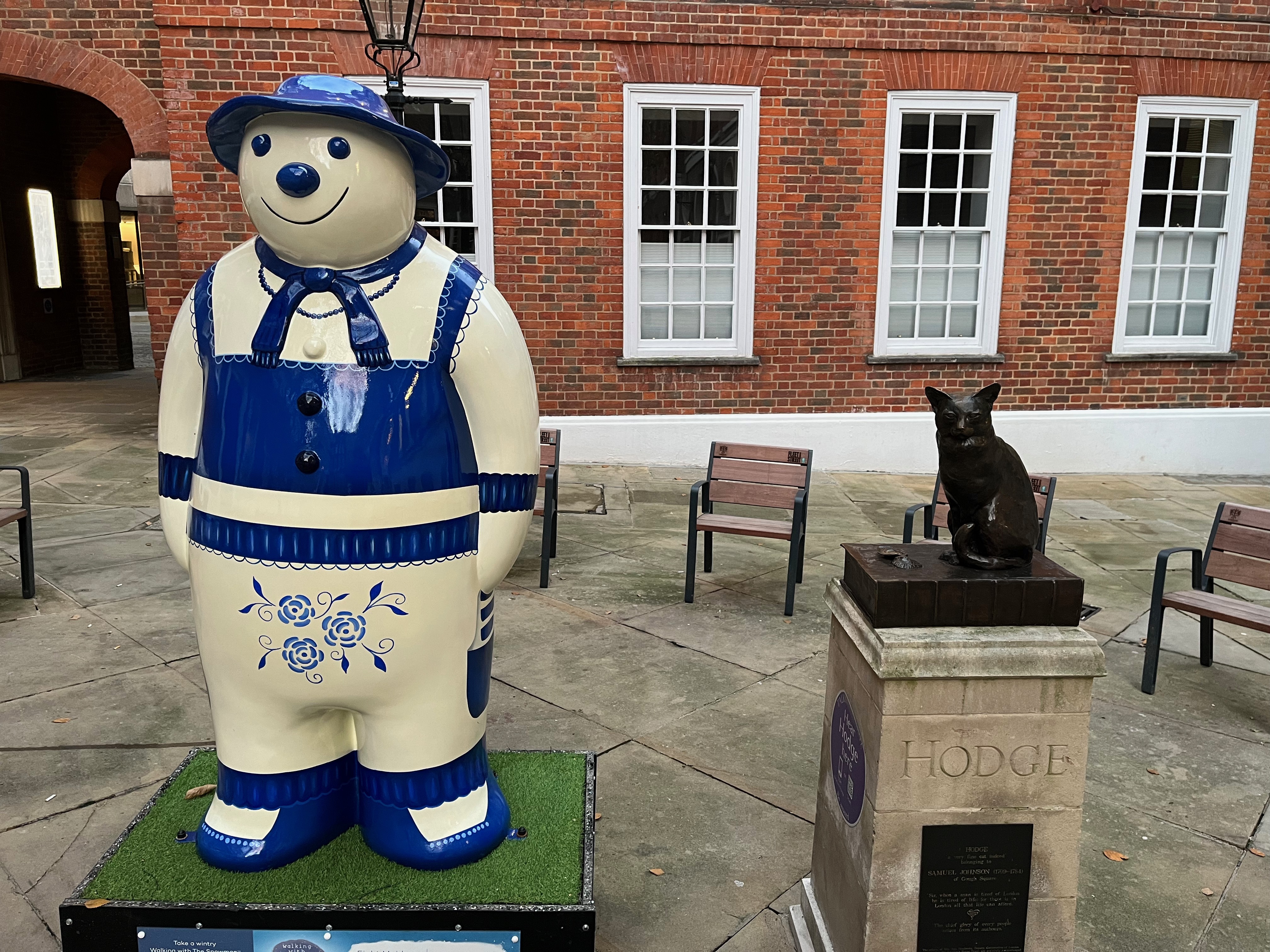 Sculpture of The Snowman called Eight Maids a-Milking, wearing a maid's apron, shoes, scarf and hat that are all dark blue in colour, in contrast to his white body. A few flowers are also painted in dark blue on his groin area. Next to the sculpture is a plinth with a small bronze statue of Hodge the cat.