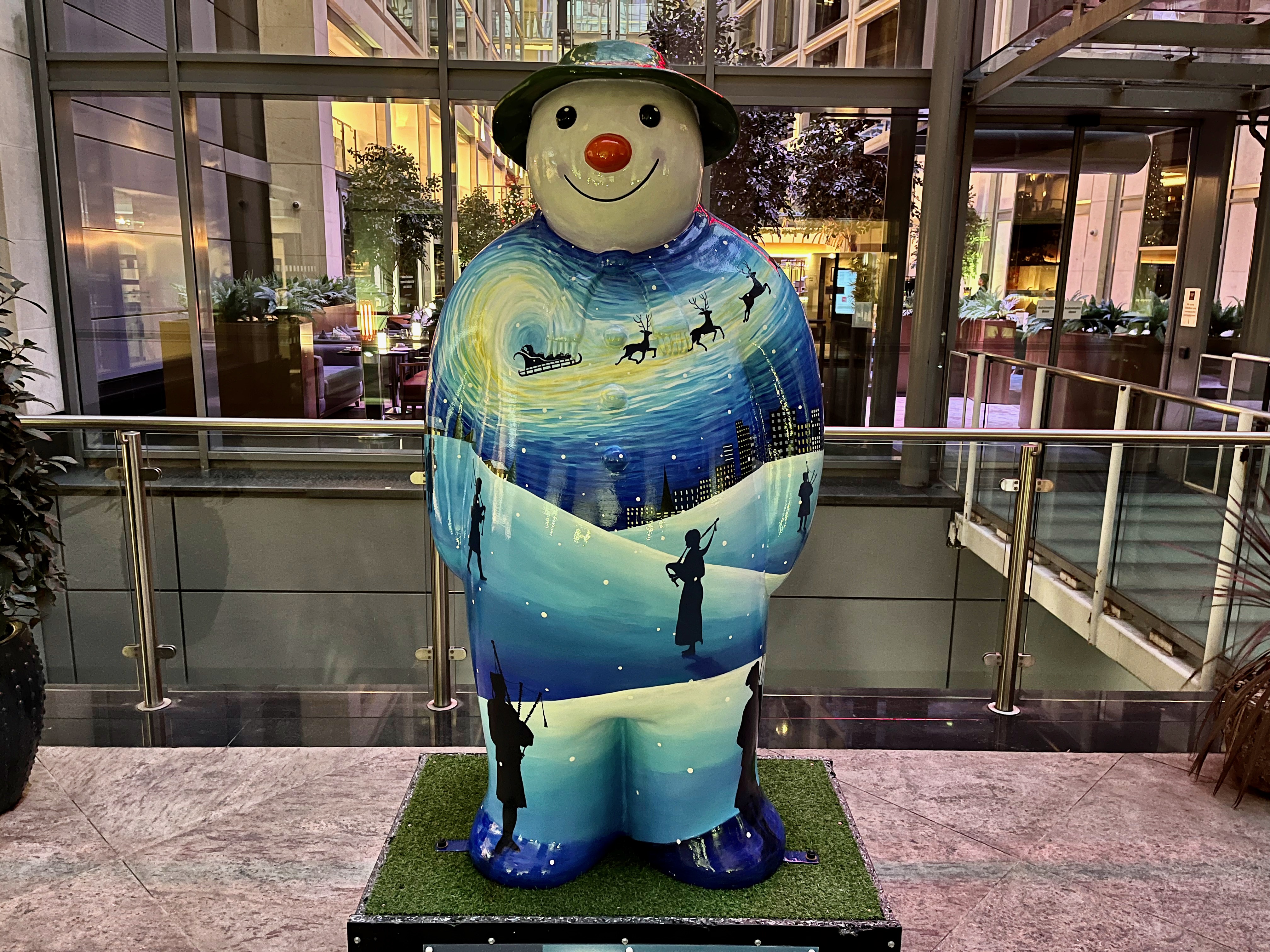Sculpture of The Snowman called Eleven Pipers Piping. It shows a landscape of snowy hills, on which silhouettes of three bagpipers are standing, while in the distance 3 reindeer pull a sleigh through the night sky, over the tops of houses and tower blocks.