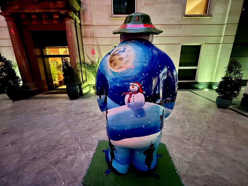 The back of the sculpture of the Snowman called Eleven Pipers Piping. It is painted to show a large round snowman, wearing a stripy scarf and a Santa hat, standing on a snowy hill in front of a starry night sky. Silhouettes of a couple of reindeer are passing across the large moon overhead.