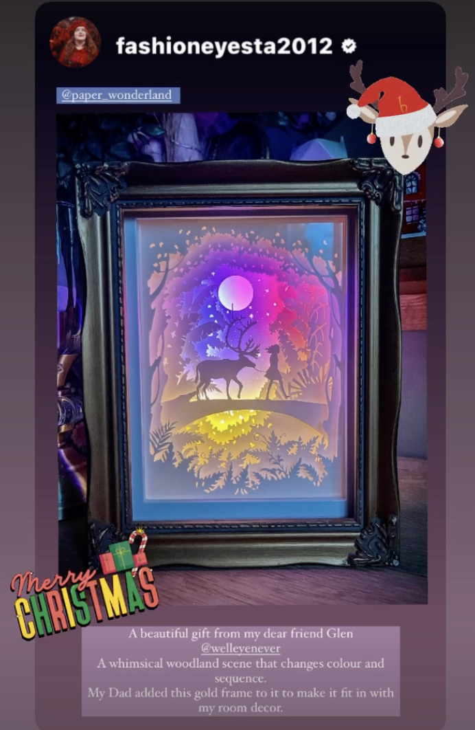Instagram post by Emily at Fashioneyesta2012, showing a framed image of a woodland scene made out of card. It shows a silhouette of deer following a girl across a short bridge between two groups of trees. Behind them, in the clearing between the trees, the moon and stars are in the sky. which is lit in rainbow colours, moving from yellow at the bottom through to dark blue and a hint of pink at the top. Text below the image reads A beautiful gift from my dear friend Glen at Well Eye Never. A whimsical woodland scene that changes colour and sequence. My Dad added this gold frame to it to make it fit in with my room decor.