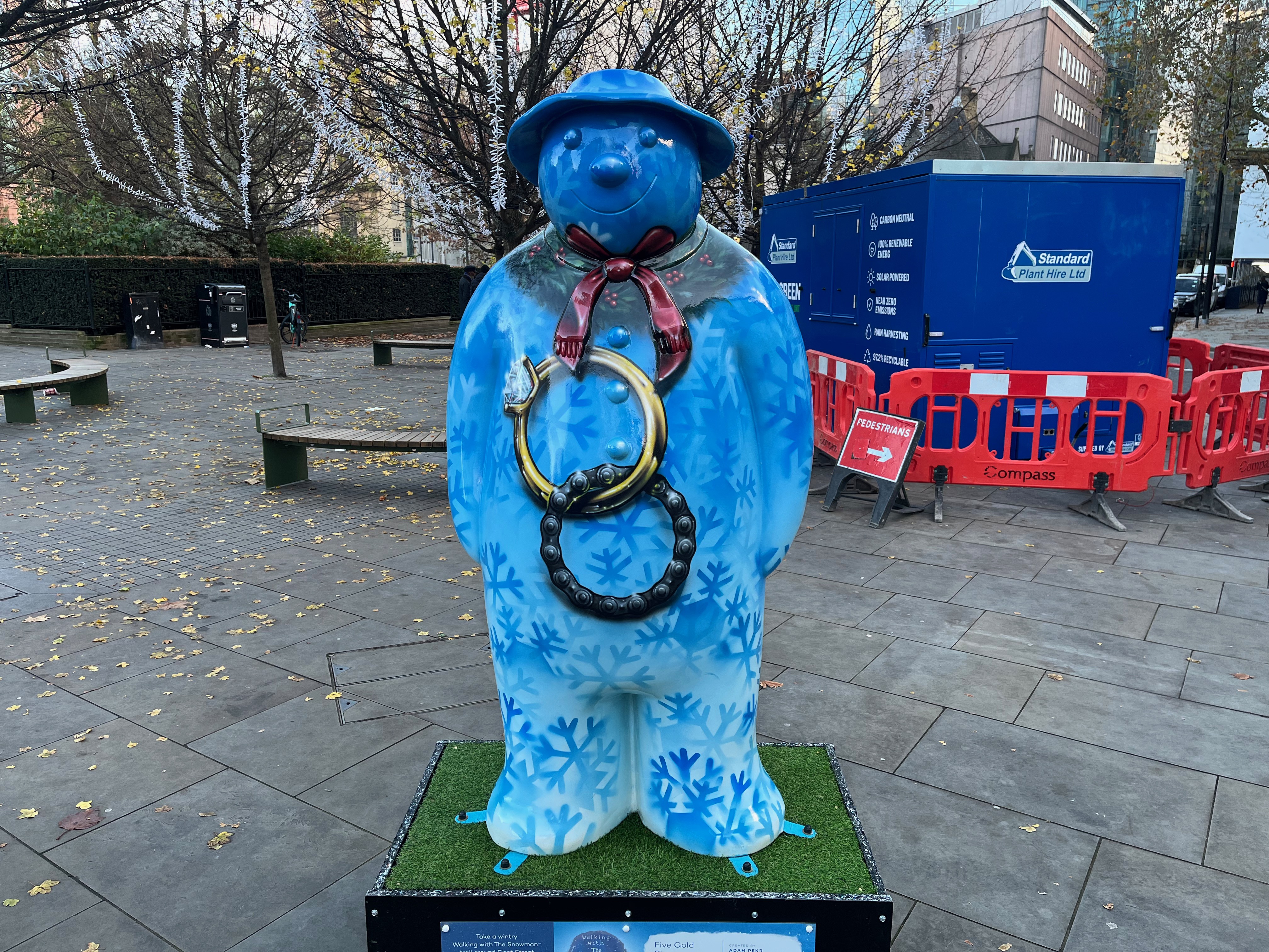 Sculpture of The Snowman called Five Gold Rings. Two large inter-linked rings, one gold with a glittering gem, and the other a dark metal, sit in the middle of his chest. The rest of his body is mostly covered in a light blue background, on which are many darker blue silhouettes of snowflakes. But there is also a strip of dark green leaves with small red berries around his neck, over which he is wearing a dark red scarf. And on top of his blue head he's wearing a blue hat.