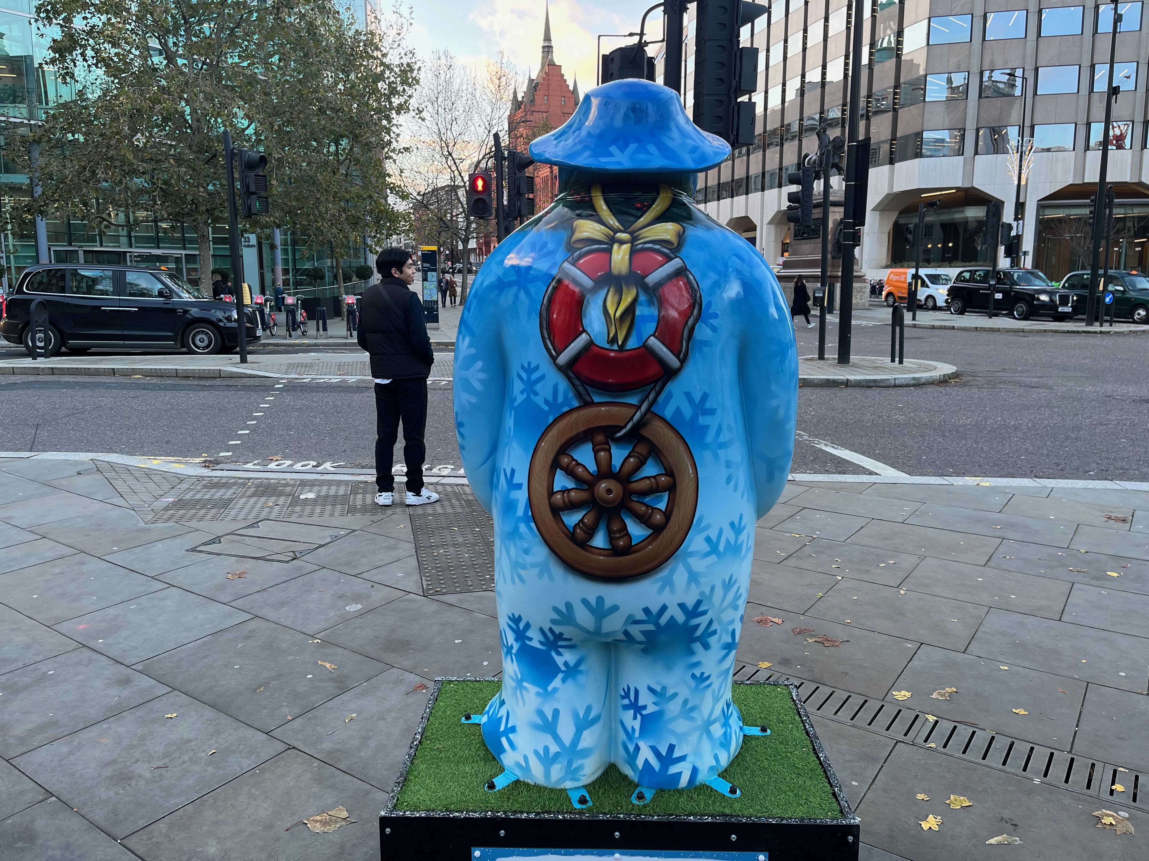 The back of the sculpture of The Snowman called Five Gold Rings. The back of his body is covered in a light blue background, on which are many darker blue silhouettes of snowflakes. On top of that, down the centre of his back, a round red life buoy ring is tied around the Snowman's neck with a yellow ribbon, while the bottom of the ring is connected by a piece of rope to a wooden wheel.