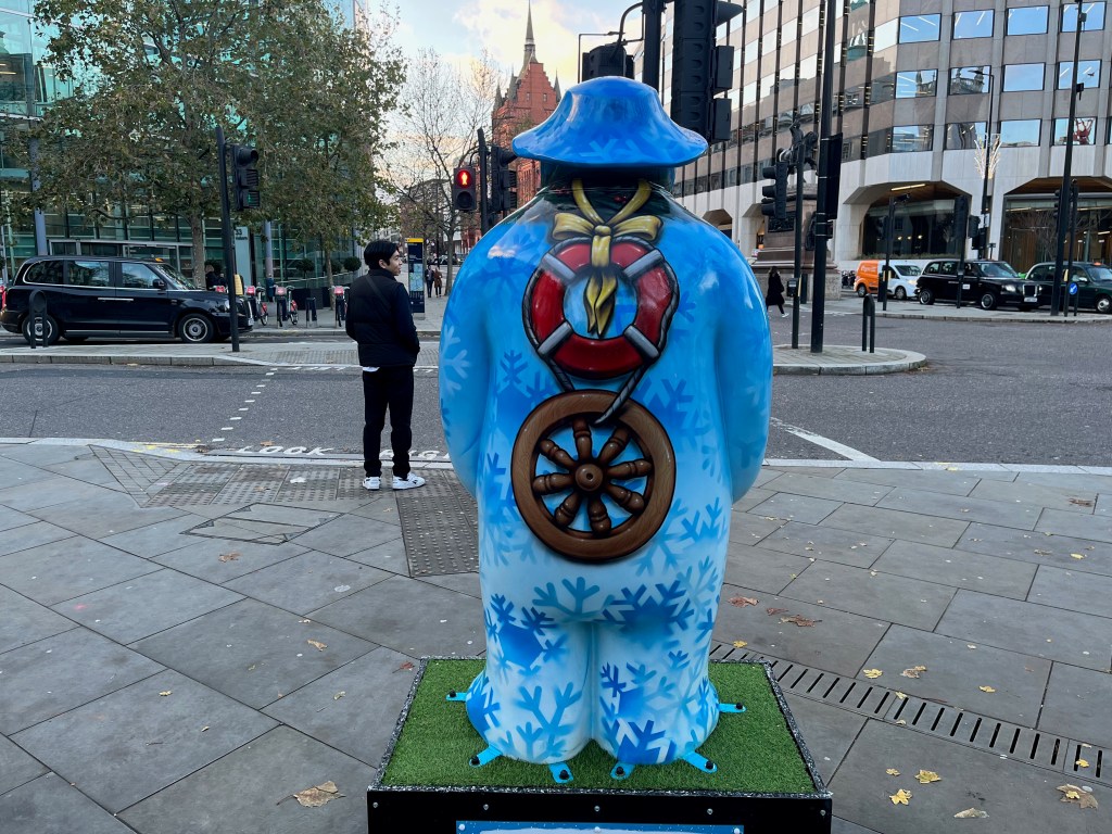 The back of the sculpture of The Snowman called Five Gold Rings. The back of his body is covered in a light blue background, on which are many darker blue silhouettes of snowflakes. On top of that, down the centre of his back, a round red life buoy ring is tied around the Snowman's neck with a yellow ribbon, while the bottom of the ring is connected by a piece of rope to a wooden wheel.