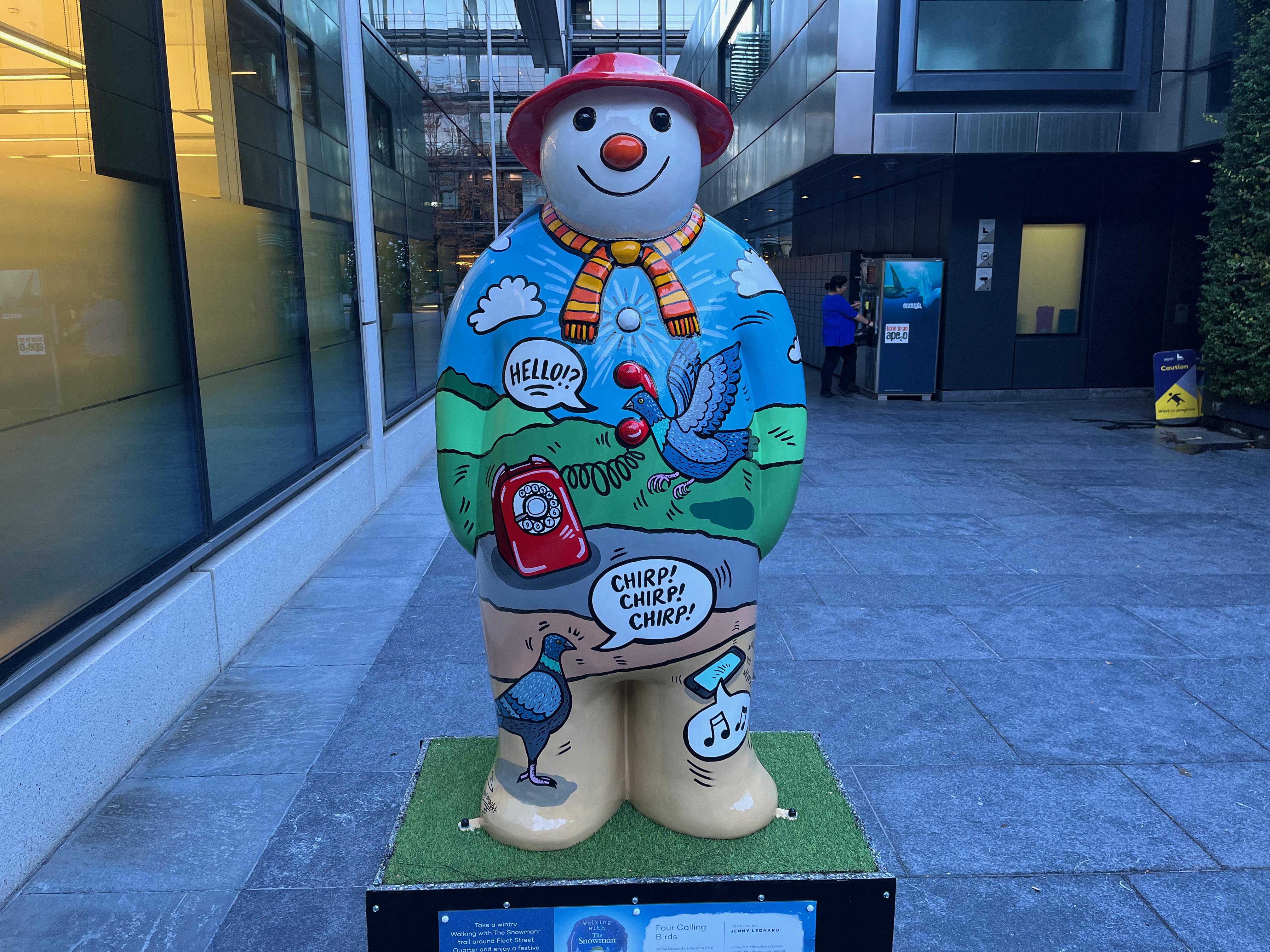 Sculpture of The Snowman called Four Calling Birds. Over a scenic background of rolling green hills against a blue sky, a bird hovers next to the floating receiver of a red rotary dial phone, next to a speech bubble saying hello, while on the Snowman's feet a bird chirps 3 times at a smartphone that has musical notes coming out of it. The Snowman is also wearing a yellow and pink striped scarf and a red hat.
