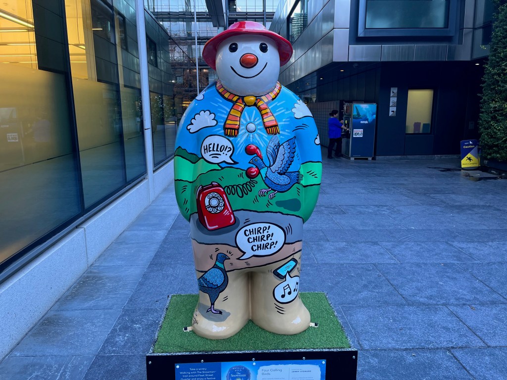 Sculpture of The Snowman called Four Calling Birds. Over a scenic background of rolling green hills against a blue sky, a bird hovers next to the floating receiver of a red rotary dial phone, next to a speech bubble saying hello, while on the Snowman's feet a bird chirps 3 times at a smartphone that has musical notes coming out of it. The Snowman is also wearing a yellow and pink striped scarf and a red hat.