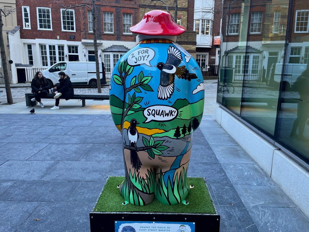 Back of a sculpture of The Snowman called Four Calling Birds. Over a scenic background of rolling green hills against a blue sky, a bird flies overhead exclaiming 2 for joy, and another bird squawks while perched on a tree branch.