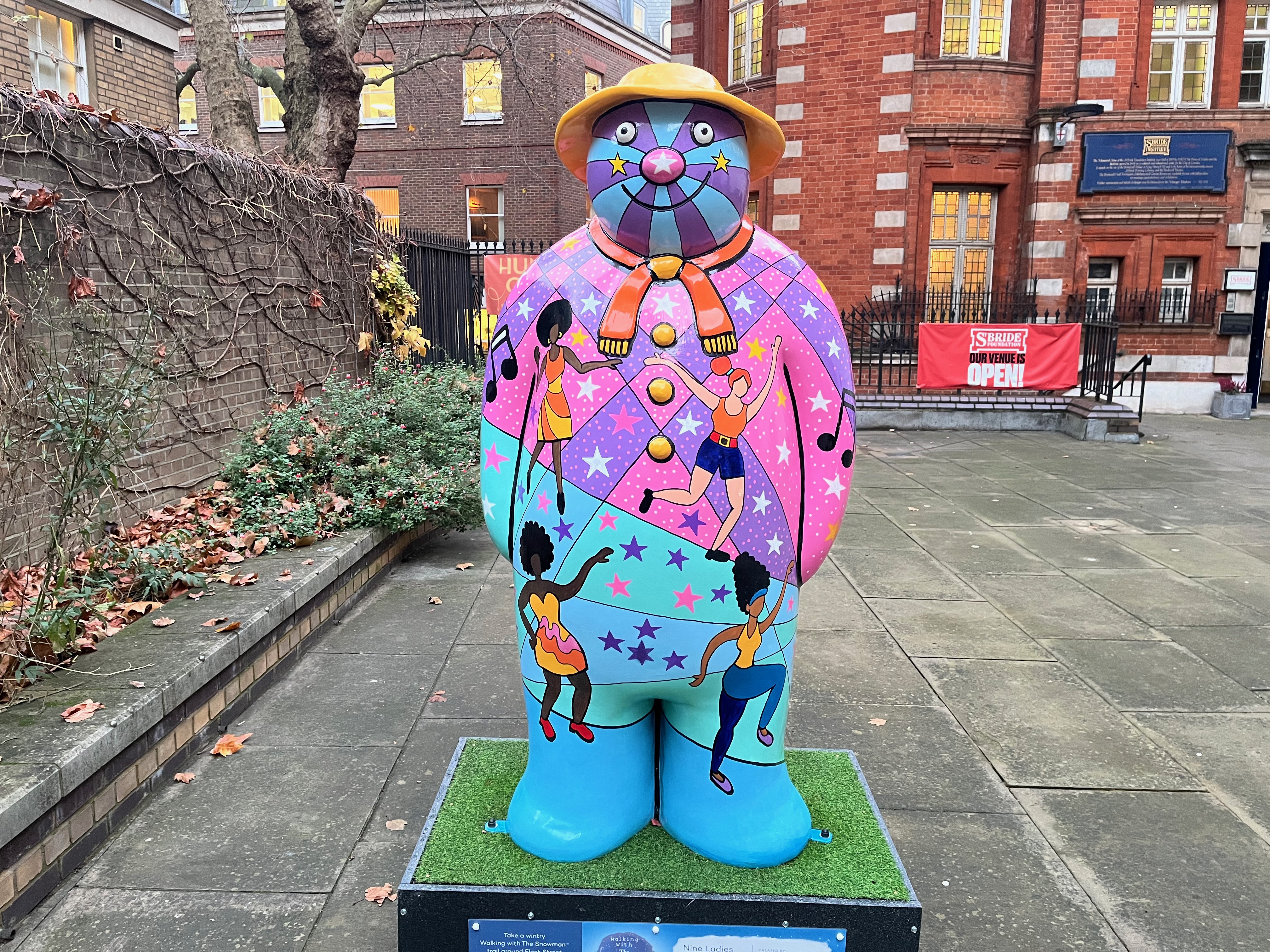 Sculpture of The Snowman called Nine Ladies Dancing. It features disco divas dancing on top of a vibrant background of colours and patterns in shades of pink, purple, blue and green, on which are stars in white, pink and blue colours. The snowman is also wearing an orange scarf and a yellow hat.