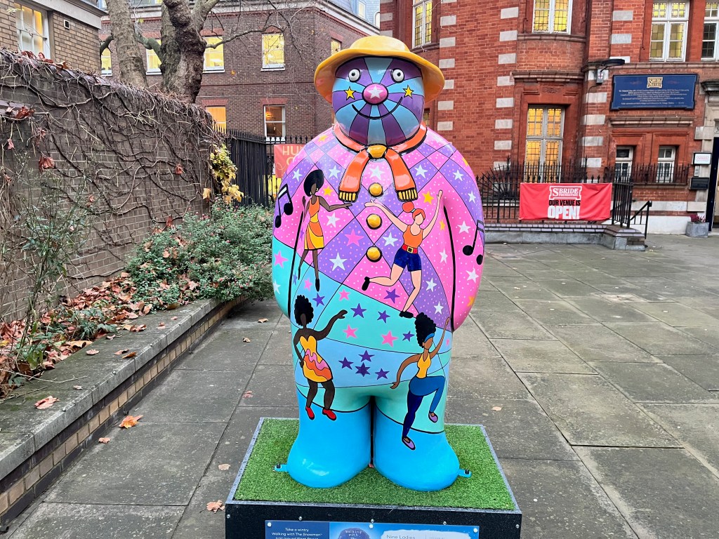 Sculpture of The Snowman called Nine Ladies Dancing. It features disco divas dancing on top of a vibrant background of colours and patterns in shades of pink, purple, blue and green, on which are stars in white, pink and blue colours. The snowman is also wearing an orange scarf and a yellow hat.
