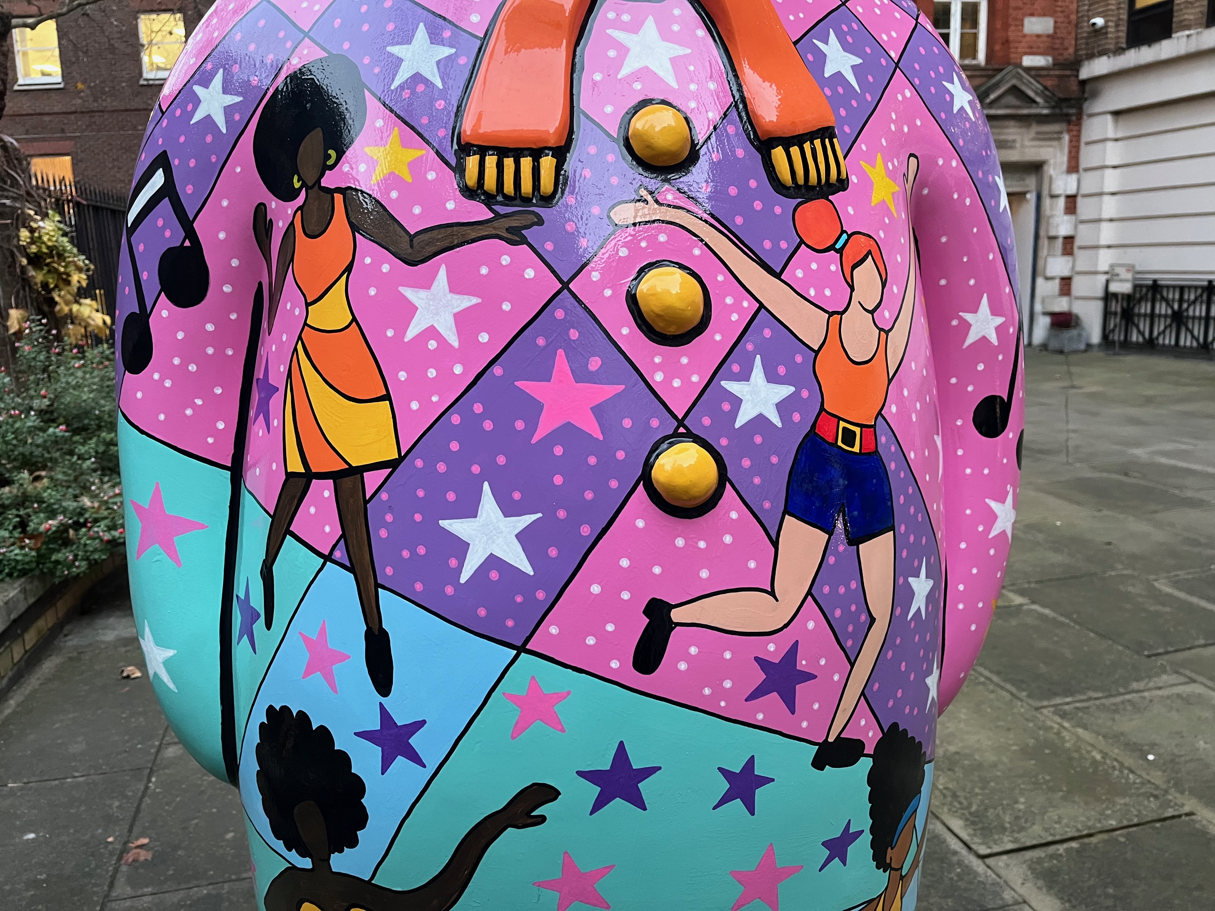 Close-up of 2 ladies dancing on the sculpture of The Snowman called Nine Ladies dancing. One is a black lady with a large black hairdo, wearing an orange and yellow striped sleeveless dress, while the other is a white redheaded lady wearing an orange sleeveless top and dark blue shorts with a red belt.
