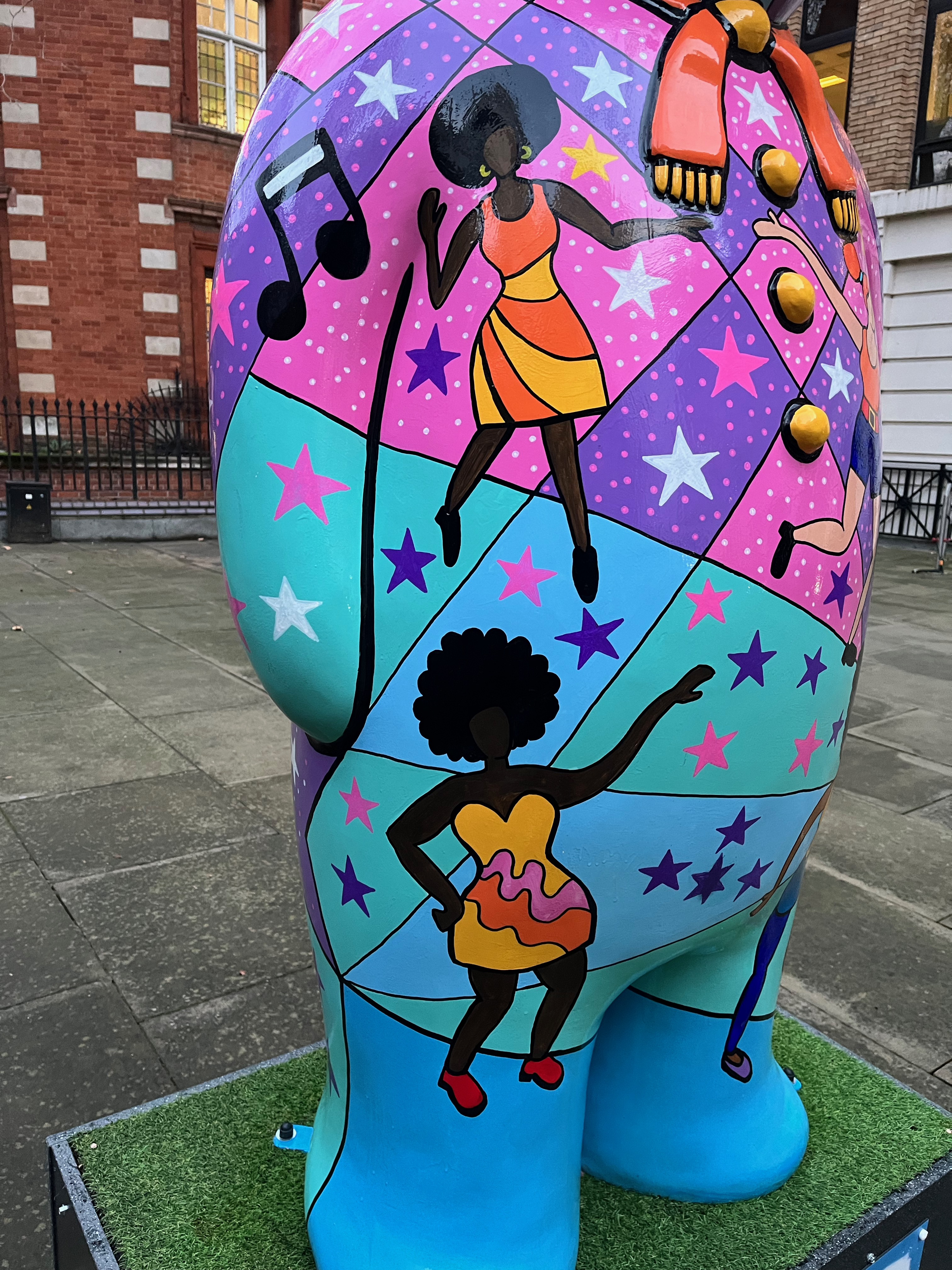 Close-up of 2 ladies dancing on the side of the Snowman sculpture called Nine Ladies dancing. One is a black lady with a large black hairdo, wearing an orange and yellow striped sleeveless dress, while below her the other is a black lady wearing a yellow strapless dress with wavy pink and red stripes 