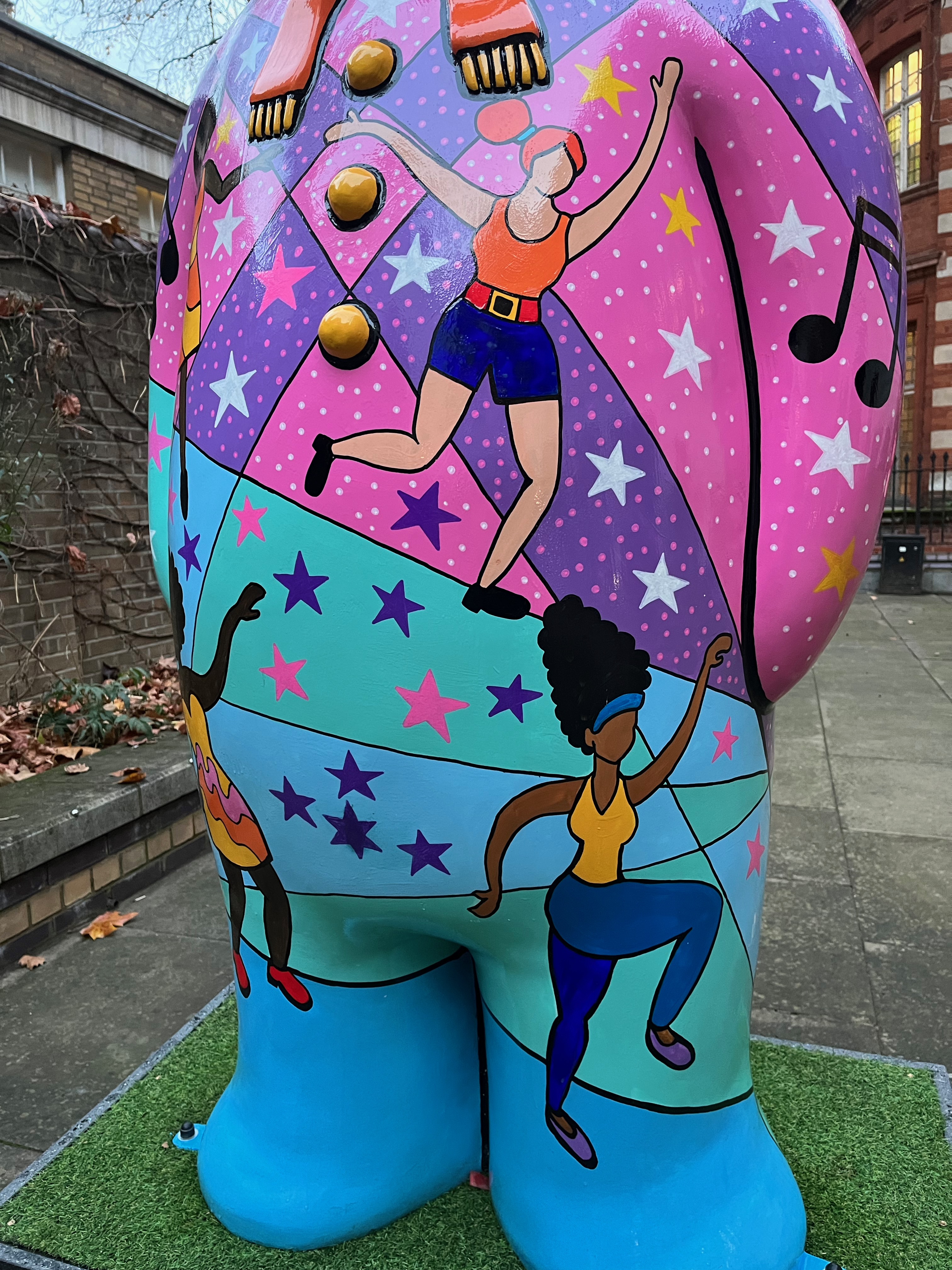Close-up of 2 ladies dancing on the side of the Snowman sculpture called Nine Ladies dancing. One is a white redheaded lady wearing an orange sleeveless top and dark blue shorts with a red belt, while below her the other is a black lady sporting a tall black hairdo with a blue bandana, and wearing blue trousers with a sleeveless yellow top