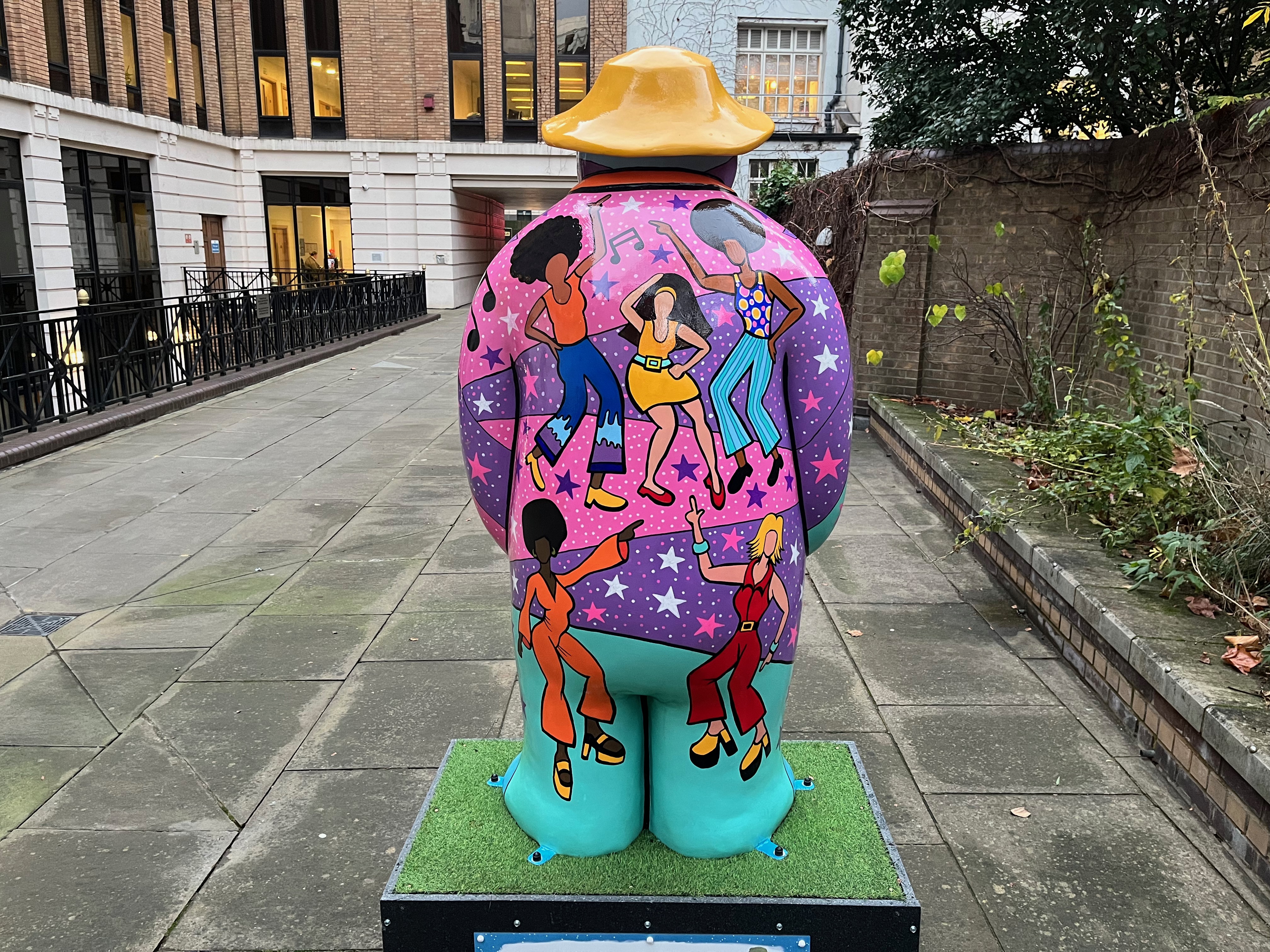 The back of the sculpture of The Snowman called Nine Ladies Dancing. It features disco divas dancing on top of a vibrant background of colours and patterns in shades of pink, purple, blue and green, on which are stars in white, pink and blue colours, lots of white dots to represent points of light generated by a mirrorball, and some black musical notes.