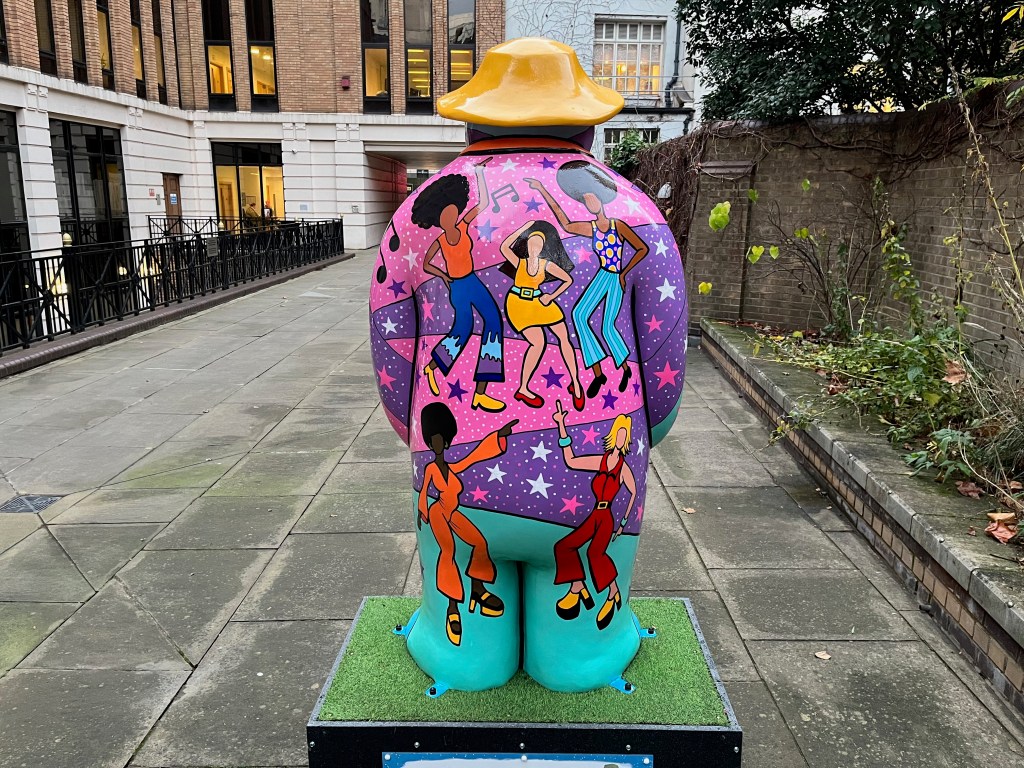 The back of the sculpture of The Snowman called Nine Ladies Dancing. It features disco divas dancing on top of a vibrant background of colours and patterns in shades of pink, purple, blue and green, on which are stars in white, pink and blue colours, lots of white dots to represent points of light generated by a mirrorball, and some black musical notes.