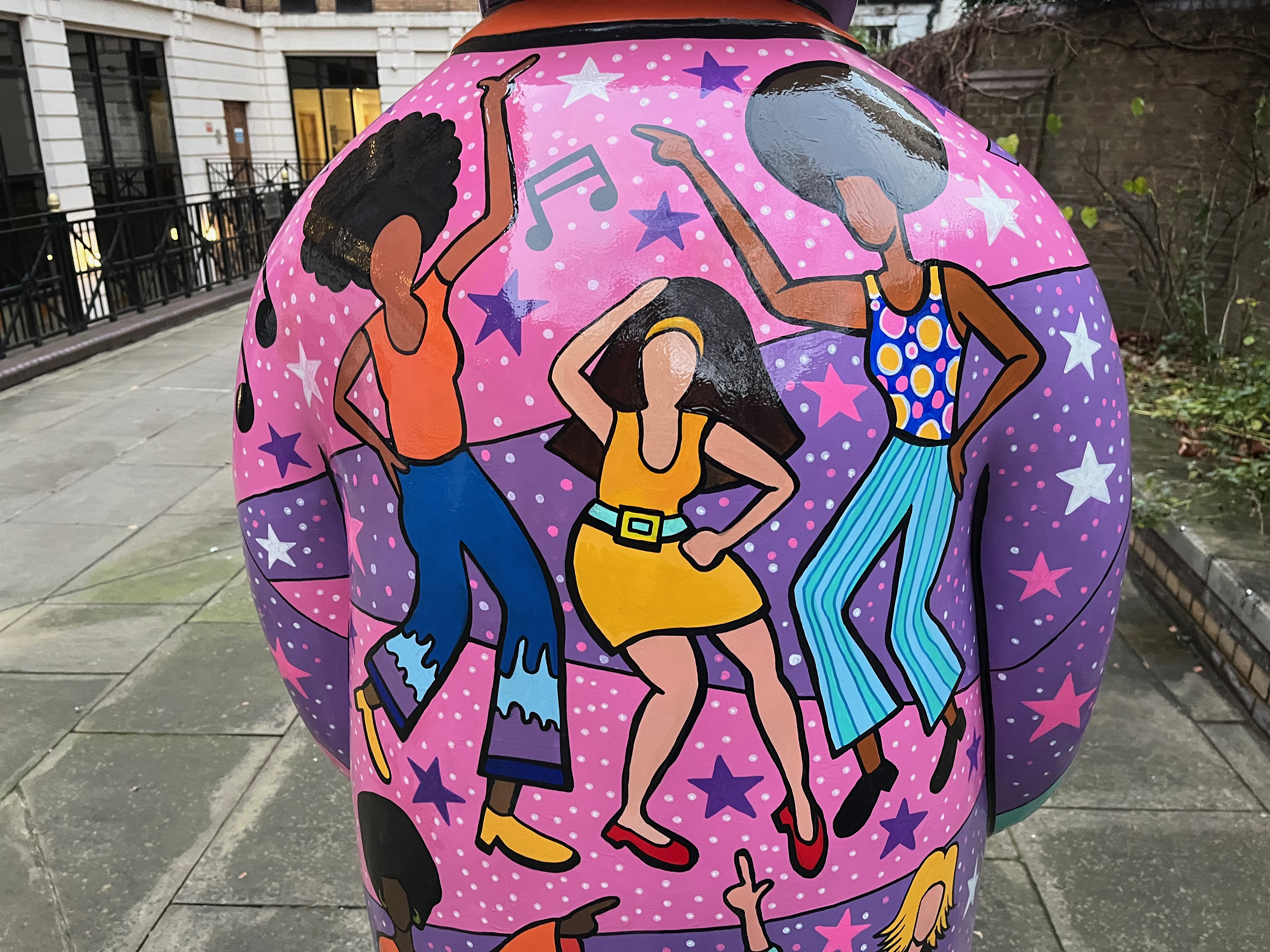 Close-up of 3 ladies dancing on the sculpture of The Snowman called Nine Ladies Dancing. One is a black lady with a large black hairdo, wearing an orange sleeveless top and blue trousers, the second is a white lady with long black hair wearing an orange dress with a white belt, and the third is a black lady with large black hair wearing light blue trousers and a blue sleeveless top with large yellow circles and small pink circles all over it.