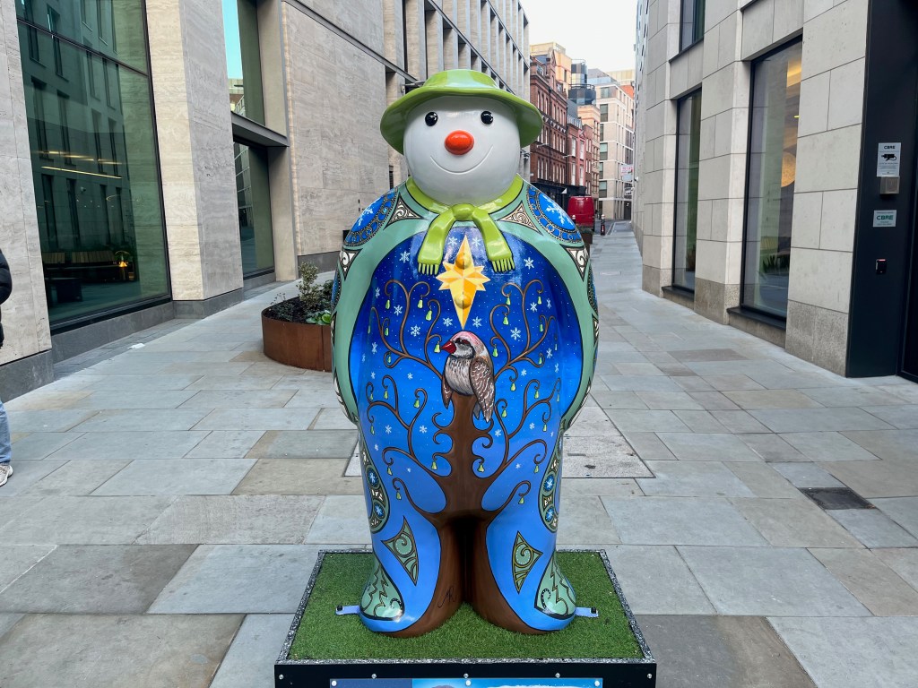Sculpture of The Snowman called Partridge In A Pear Tree. The front of his body, from his chest down to his feet, is painted to show a partridge sitting in a tall tree that has small pears hanging from its curling branches. There is a big bright yellow star just above the partridge, and there are white snowflakes falling in the starry night sky in the background. The Snowman's smiling face is white with a red nose, and he's wearing a green hat and a screen scarf.