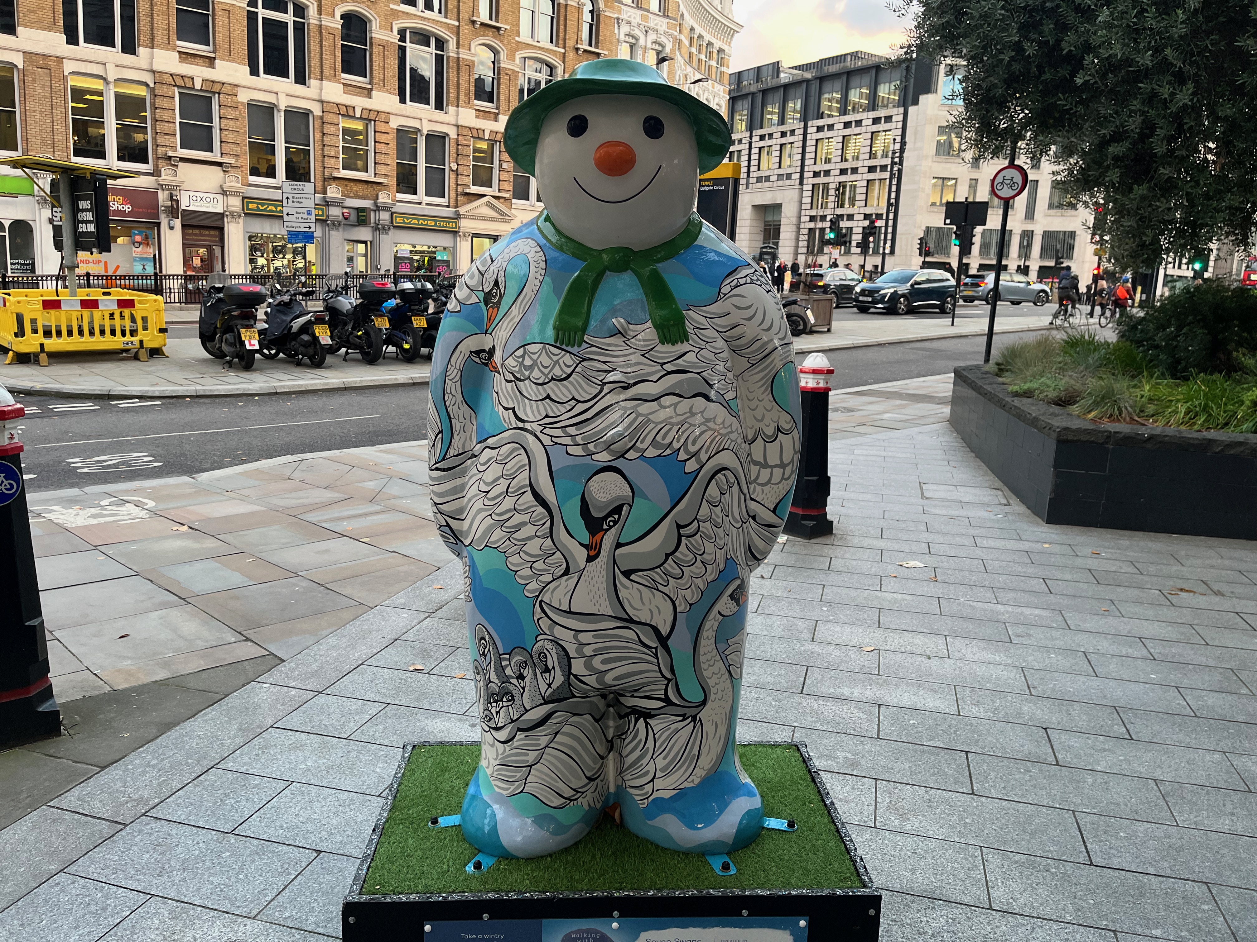 Sculpture of The Snowman called Seven Swans a-Swimming. One large swan dominates the centre of the Snowman's body, with the large wings of other swans protruding into view from the sides, and there are also some baby swans visible at the top of his right leg, wrapped up in a larger swan's feathered wing. The Snowman is also wearing a green scarf and a green hat.