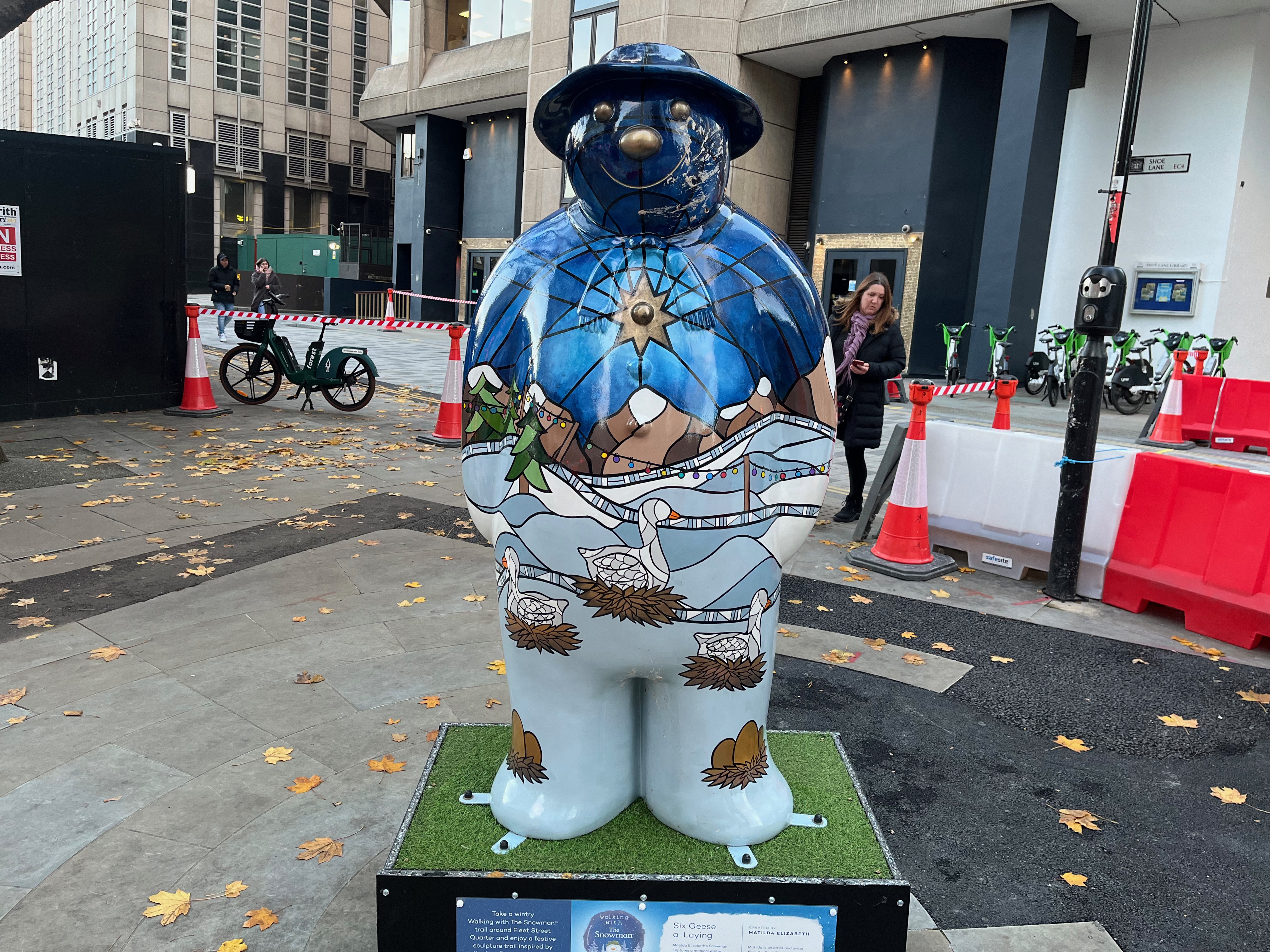 Sculpture of The Snowman called Six Geese a-Laying. His body is painted in a style a bit like stained glass, with geese sitting on nests in front of a snowy landscape with snow-topped mountains in the background, beneath a golden star in the sky. Each of his feet also has a nest containing a few eggs. His face and hat are a dark blue colour, with his eyes, nose and mouth standing out in gold.
