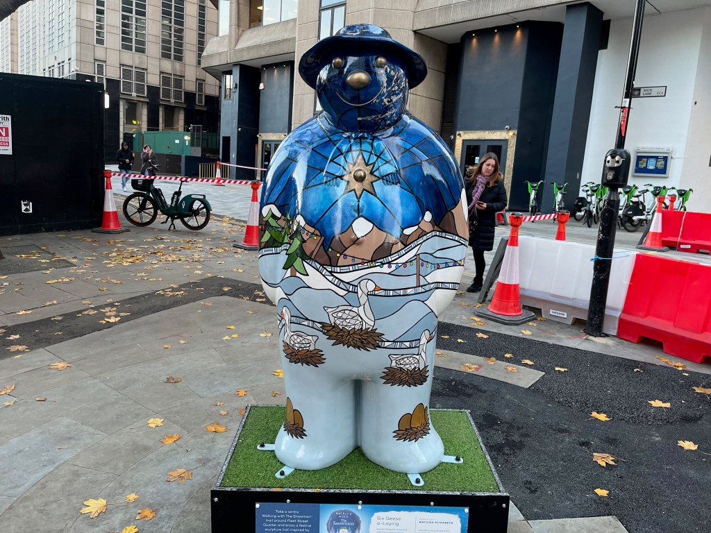 Sculpture of The Snowman called Six Geese a-Laying. His body is painted in a style a bit like stained glass, with geese sitting on nests in front of a snowy landscape with snow-topped mountains in the background, beneath a golden star in the sky. Each of his feet also has a nest containing a few eggs. His face and hat are a dark blue colour, with his eyes, nose and mouth standing out in gold.