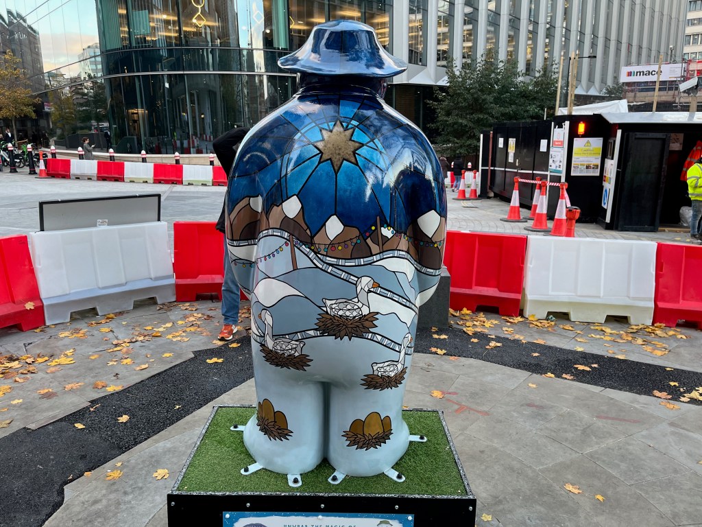 Back of the sculpture of The Snowman called Six Geese a-Laying. His back is painted in a style a bit like stained glass, with geese sitting on nests in front of a snowy landscape with snow-topped mountains in the background, beneath a golden star in the sky. Each of his feet also has a nest containing a few eggs.
