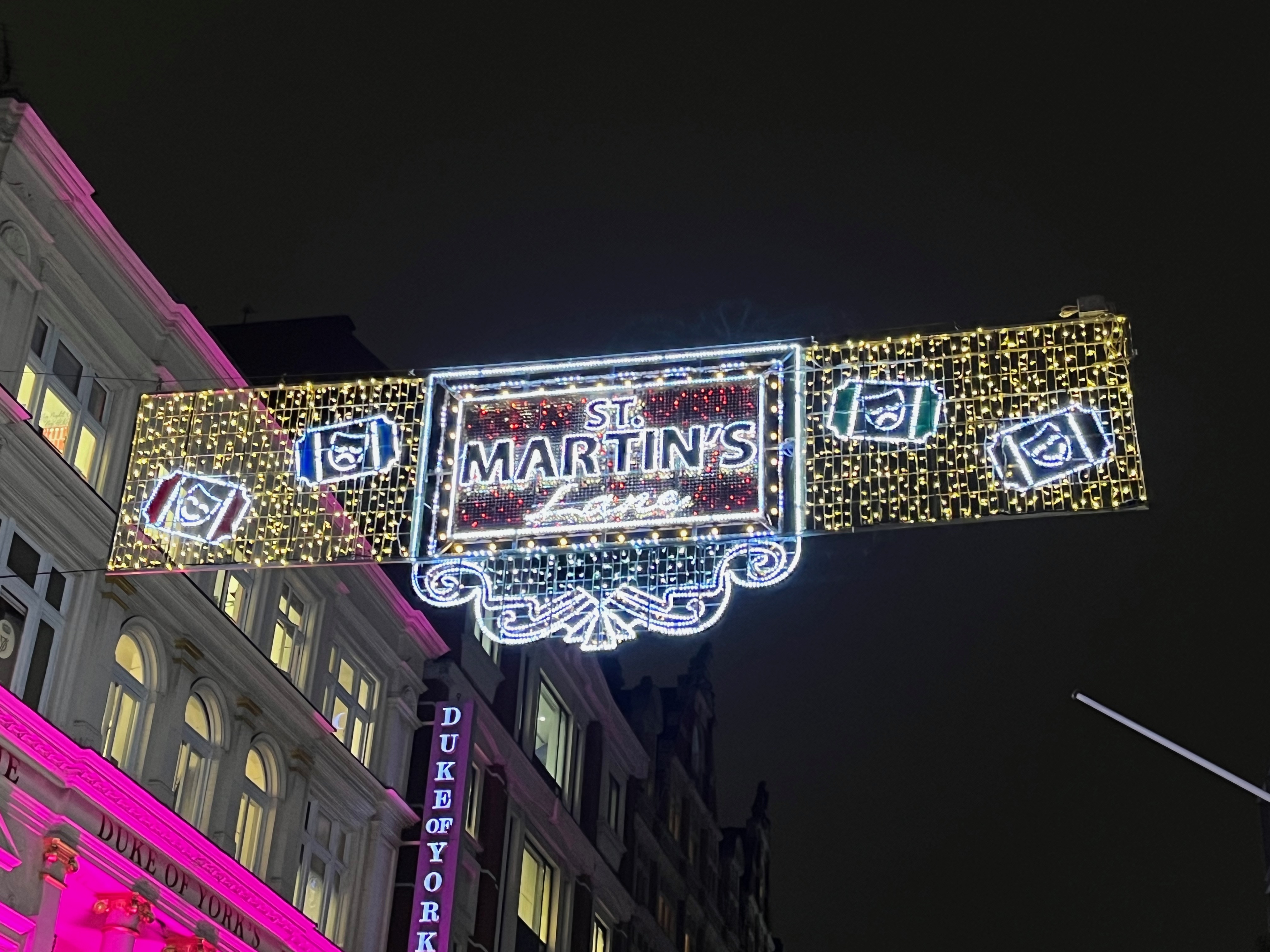 A lit up Christmas sign above St Martin's Lane, featuring the name of the street in the centre, ornate framing beneath it, and images of smiling and sad faces either side.