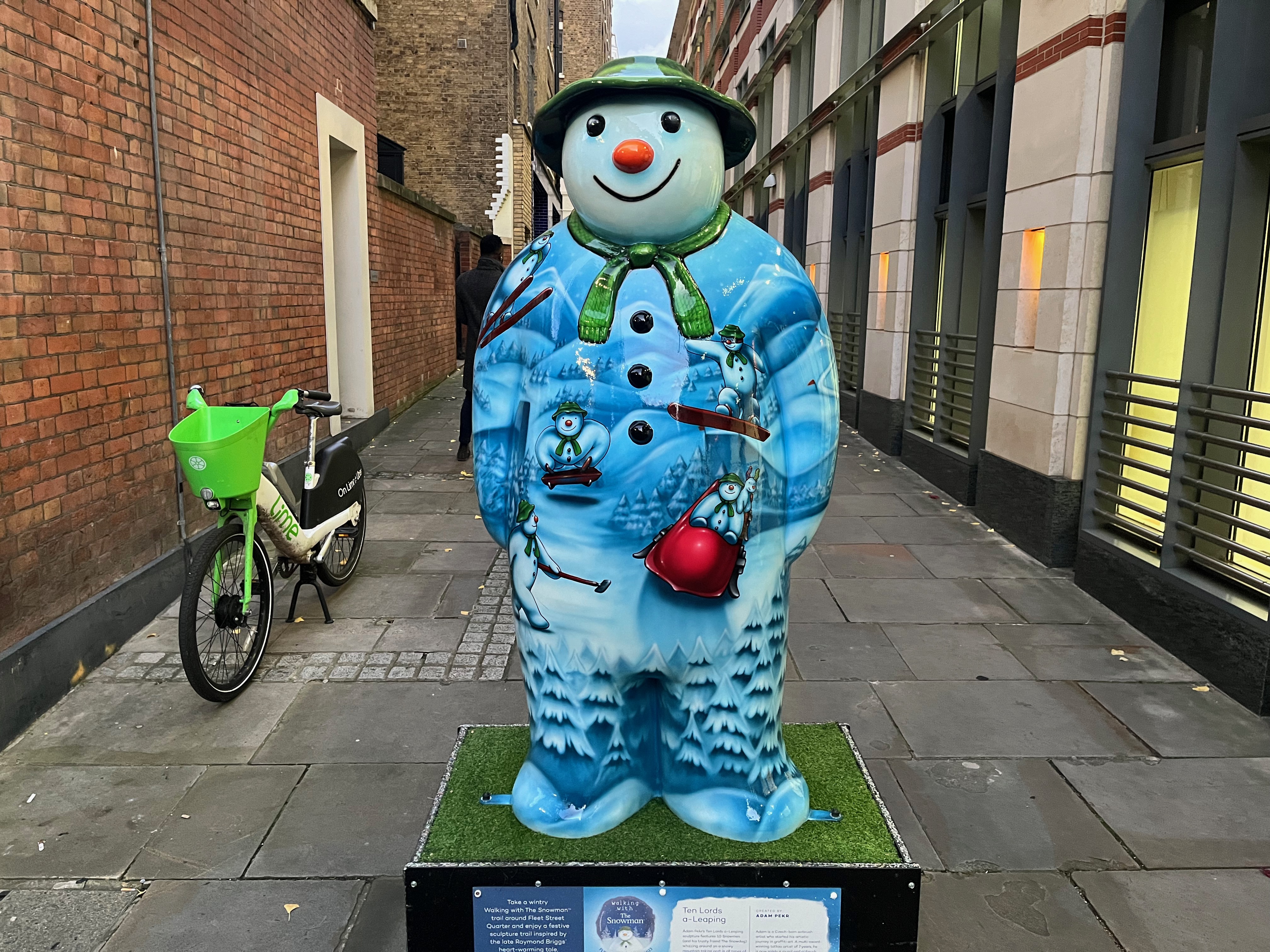 Sculpture of The Snowman called Ten Lords a-Leaping. HIs body is covered in a snowy landscape scene, on which there are lots of images of the Snowman engaging in various snow-related activities, including sledging and skiing. Just like those small drawings on him, the sculpture of the Snowman is wearing a green scarf and a green hat, framing his smiling white face.
