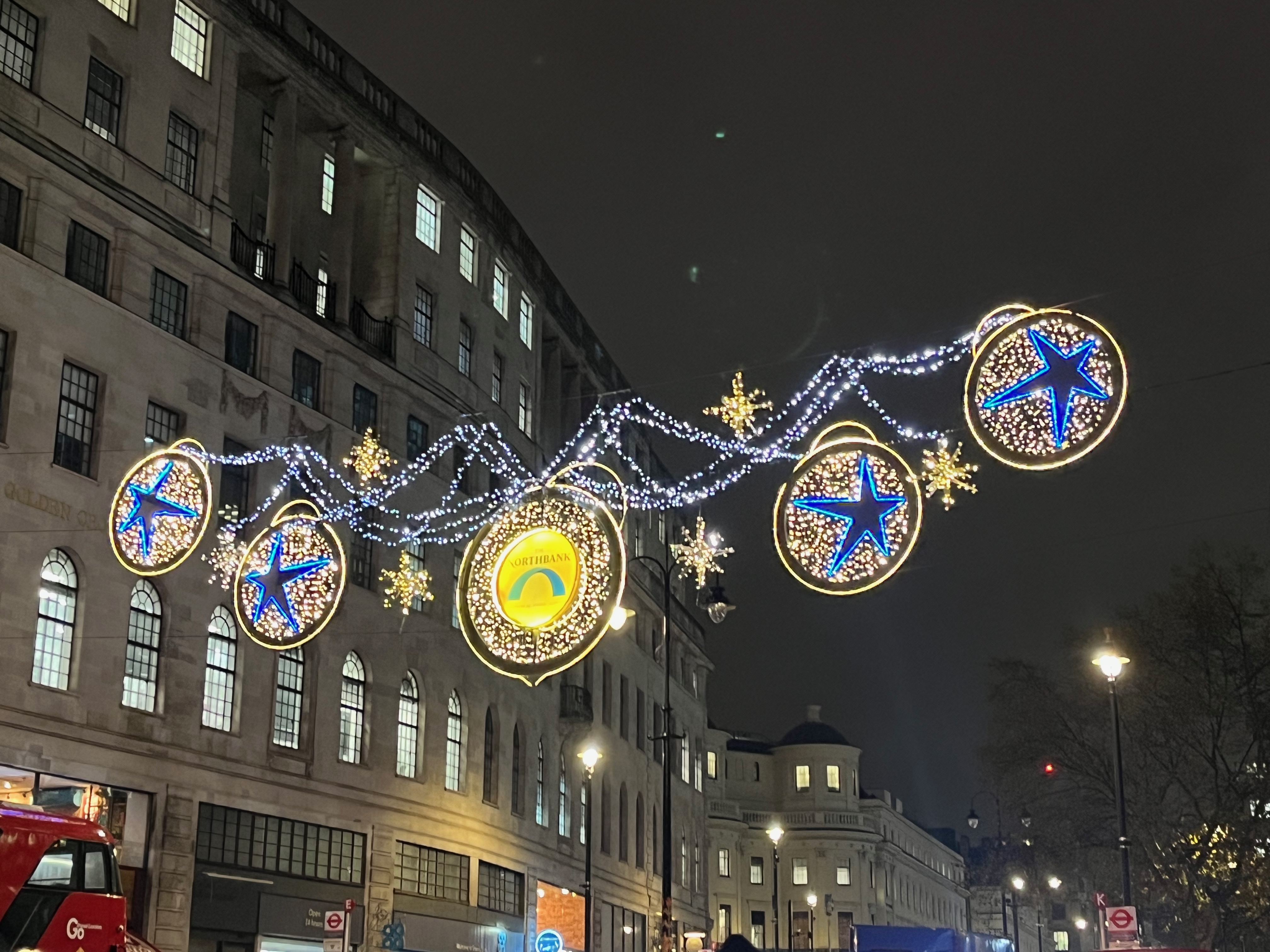 Christmas lights above the Strand, made up of 5 discs lit up in gold, connected by strings lit up in white. The central disc has a yellow centre, containing the word Northbank above a blue arch. The other 4 discs - two either side of the central one - each contain a blue star, slightly stretched and offset from its centre.