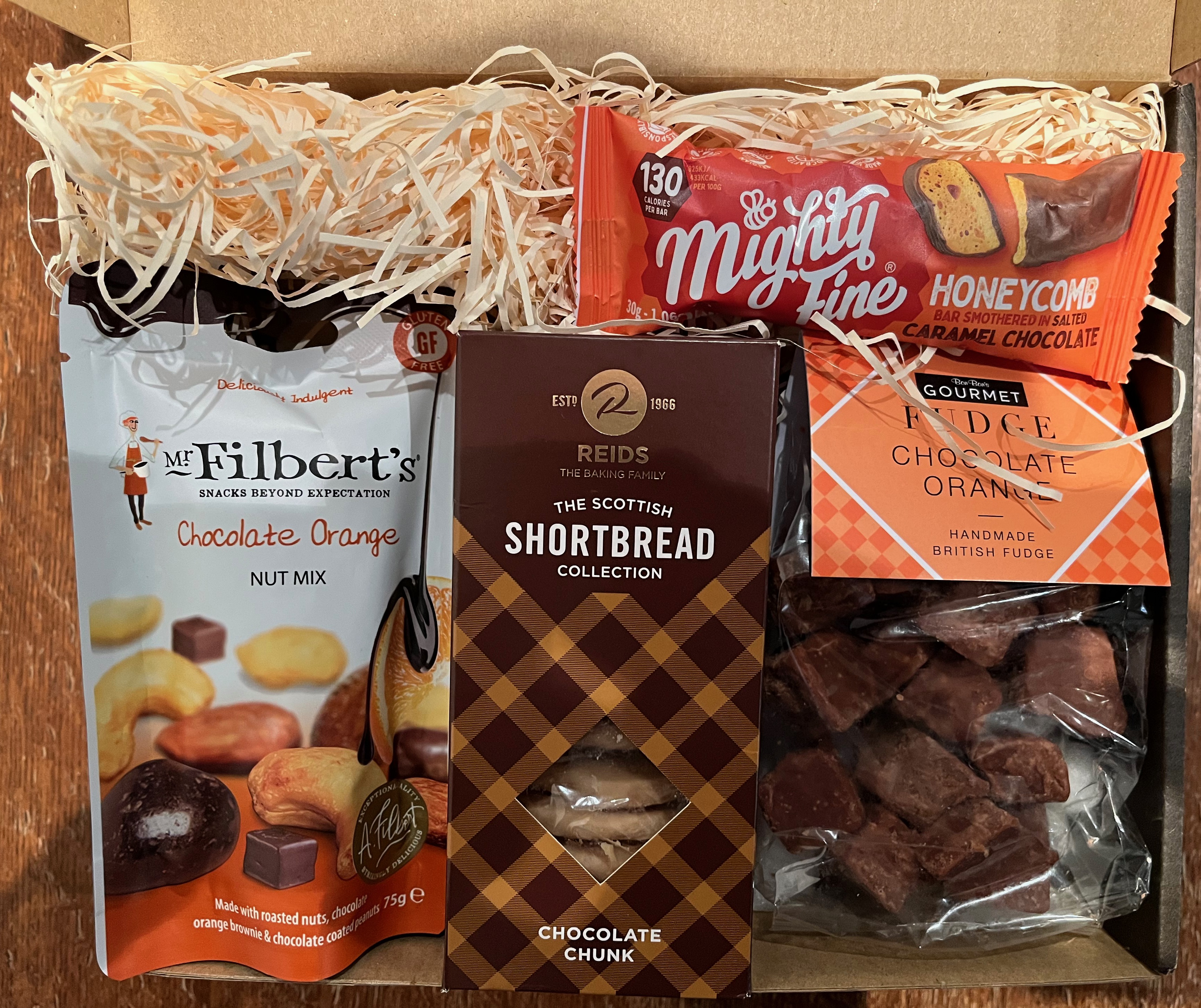 Various chocolate treats in a box lined with straw-like shavings of paper. The goodies include Mr Filbert's Chocolate Orange Nut Mix, Reids Scottish Shortbread Selection, Mighty Fine Honeycomb Bar covered in caramel chocolate, and Bon Bon's Gourmet Chocolate Orange Fudge.