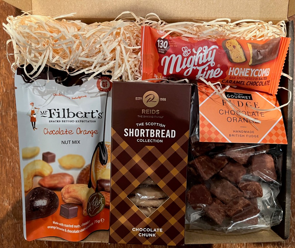 Various chocolate treats in a box lined with straw-like shavings of paper. The goodies include Mr Filbert's Chocolate Orange Nut Mix, Reids Scottish Shortbread Selection, Mighty Fine Honeycomb Bar covered in caramel chocolate, and Bon Bon's Gourmet Chocolate Orange Fudge.