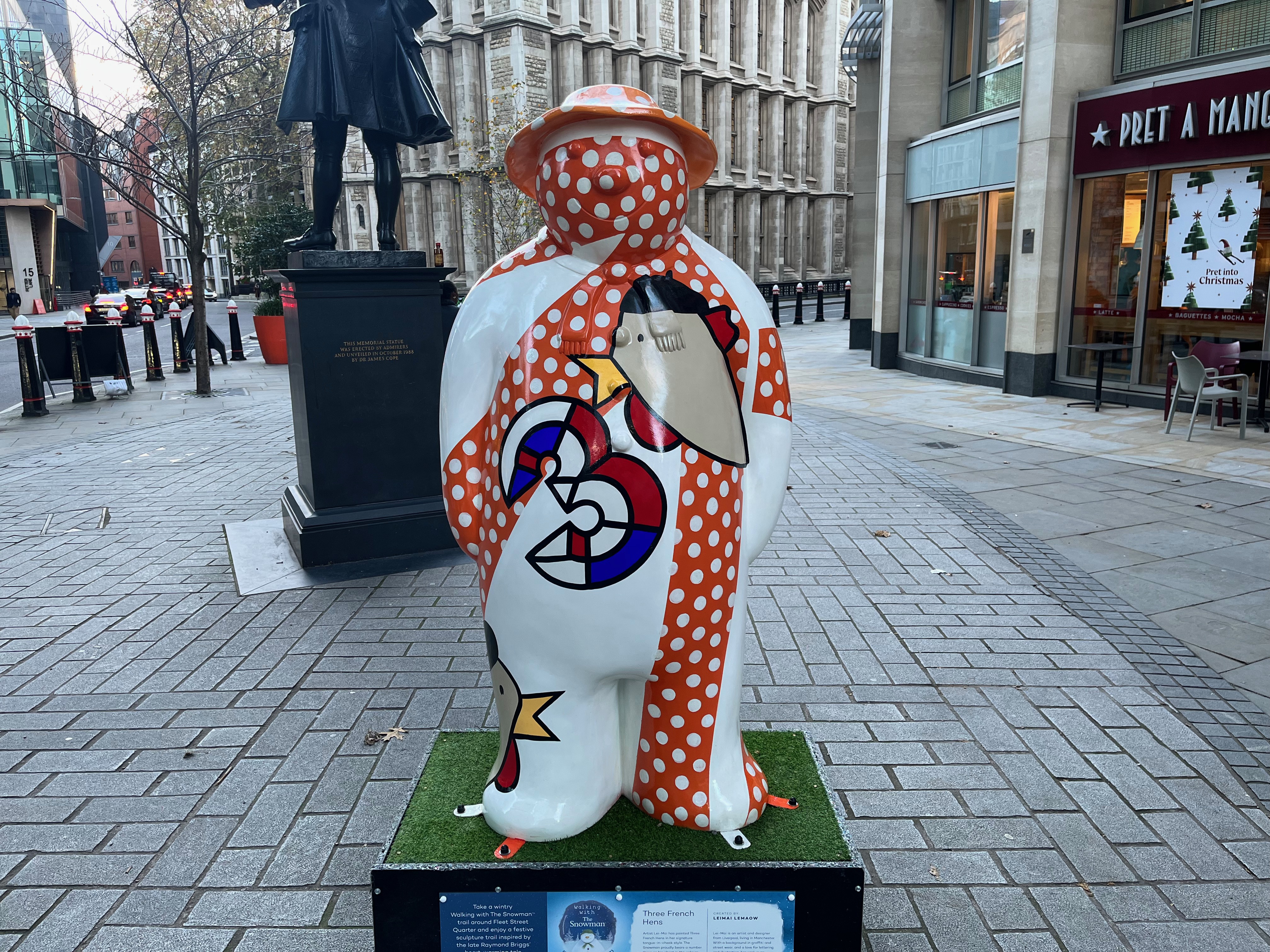 Sculpture of The Snowman called Three French Hens. His body is mainly white, but there are a few thick bands of red with white polka dots, one stretching from his left foot up to his left shoulder, another going diagonally from his right hand up to his left shoulder, and another covering his face. Over the top of these bands on his chest is a figure of a hen made of a few abstract shapes, giving it a grey body, a small yellow beak, round red wattles, and a black comb of hair on the top. Next to that is a number 3, made of 3D segments of red, white and blue. Another hen can be seen poking its beak into view on the Snowman's right foot.