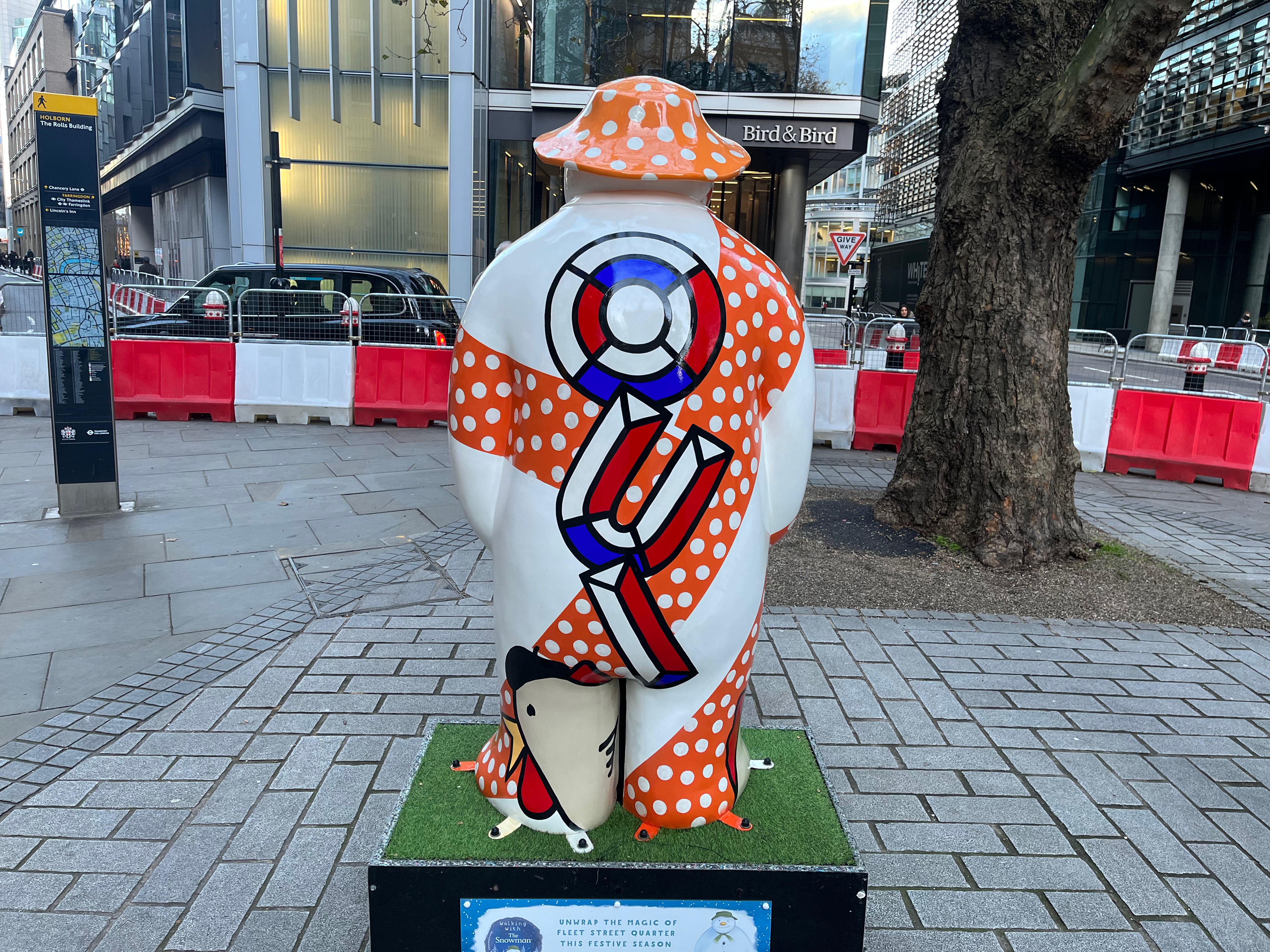 The back of the sculpture of The Snowman called Three French Hens. His body is mainly white, but there are a few thick bands of red with white polka dots wrapping around him from top to bottom and left to right. Down the centre of his back, in large letters made of 3D segments of red, white and blue, are the letters O, U and I. On the back of his left foot is a figure of a hen made of a few abstract shapes, giving it a grey body, a small yellow beak, round red wattles, and a black beret on its head.