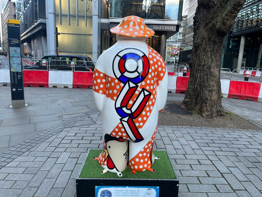 The back of the sculpture of The Snowman called Three French Hens. His body is mainly white, but there are a few thick bands of red with white polka dots wrapping around him from top to bottom and left to right. Down the centre of his back, in large letters made of 3D segments of red, white and blue, are the letters O, U and I. On the back of his left foot is a figure of a hen made of a few abstract shapes, giving it a grey body, a small yellow beak, round red wattles, and a black beret on its head.