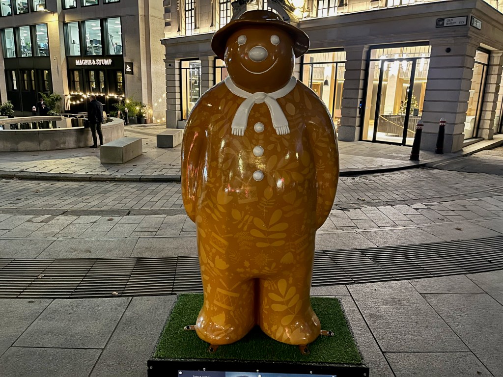 Sculpture of The Snowman called Twelve Drummers Drumming. He's painted to look like he's made of golden gingerbread, with leafy patterns in a slightly lighter shade. In contrast, the 3 buttons on his chest, his scarf and his facial features are all white, and he's wearing a red hat.