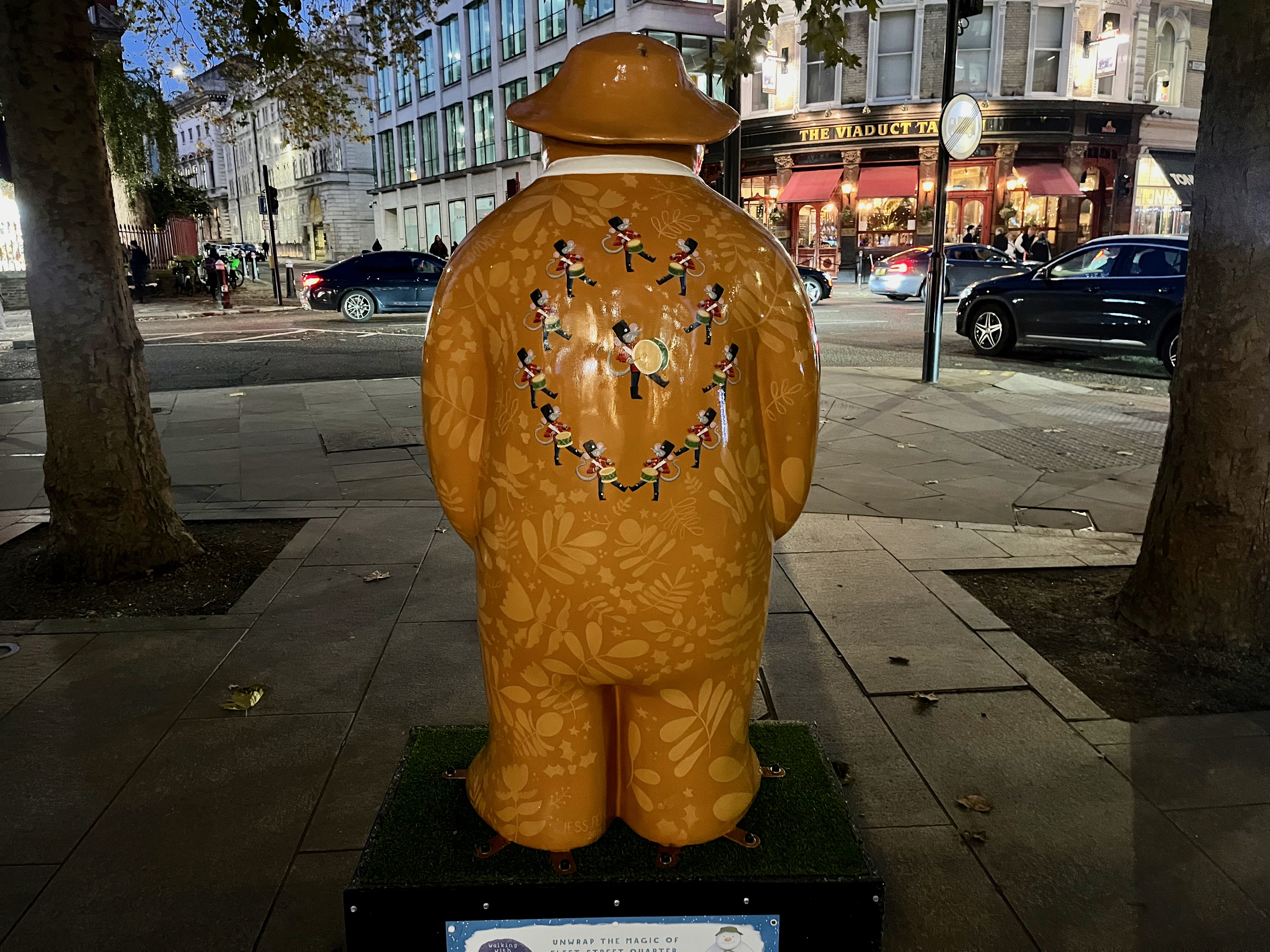 The back of the sculpture of The Snowman called Twelve Drummers Drumming. He's painted to look like he's made of golden gingerbread, with leafy patterns in a slightly lighter shade. In the centre of his back is a marching band formed of 12 small drumming mice, 11 of which are arranged in a circle with the other larger mouse in the middle.