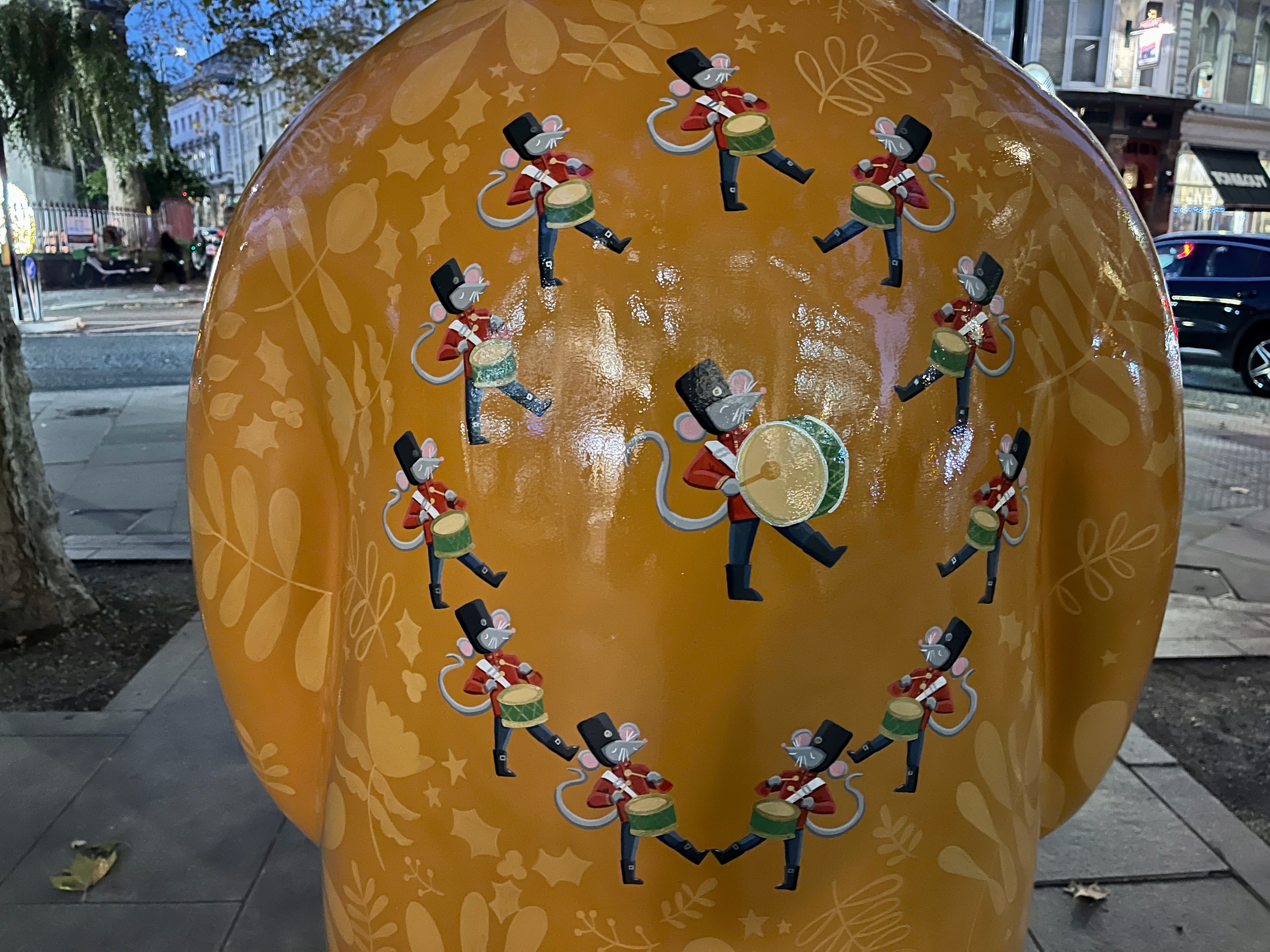 Close-up of 12 marching and drumming mice on the back of the Snowman sculpture called Twelve Drummers Drumming. 11 of the mice are forming a circle around a larger mouse in the middle. They're all dressed smartly, each wearing black trousers, a red jacket with a white diagonal sash, and a black hat.