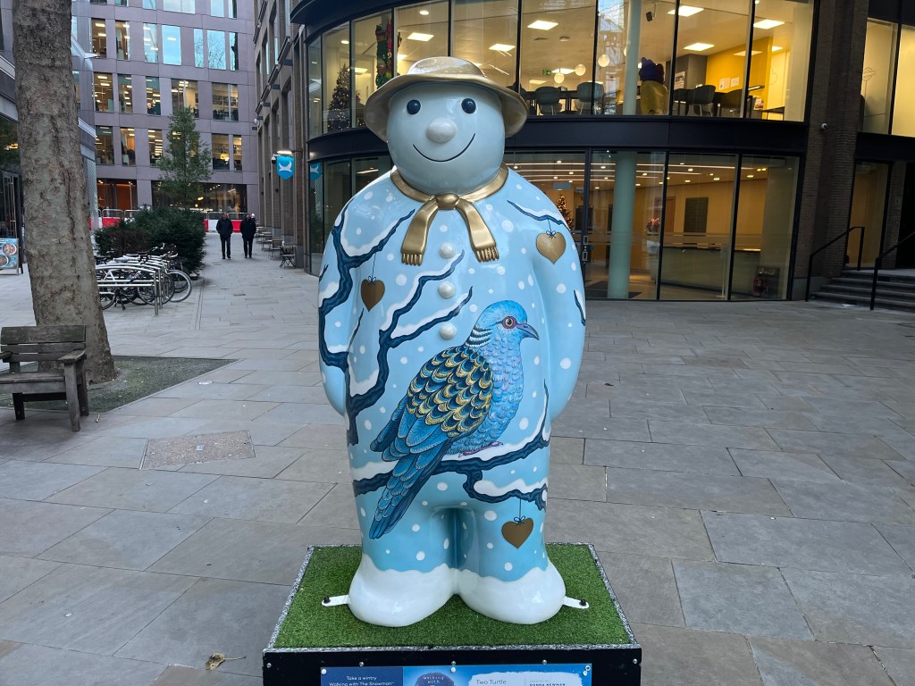 Sculpture of The Snowman called Two Turtle Doves. The front of his body shows a large blue turtle dove, with lots of feathers, sitting on a snow covered tree branch. A few red and gold coloured hearts are hanging from other branches. The Snowman is also wearing a gold scarf and a gold hat.