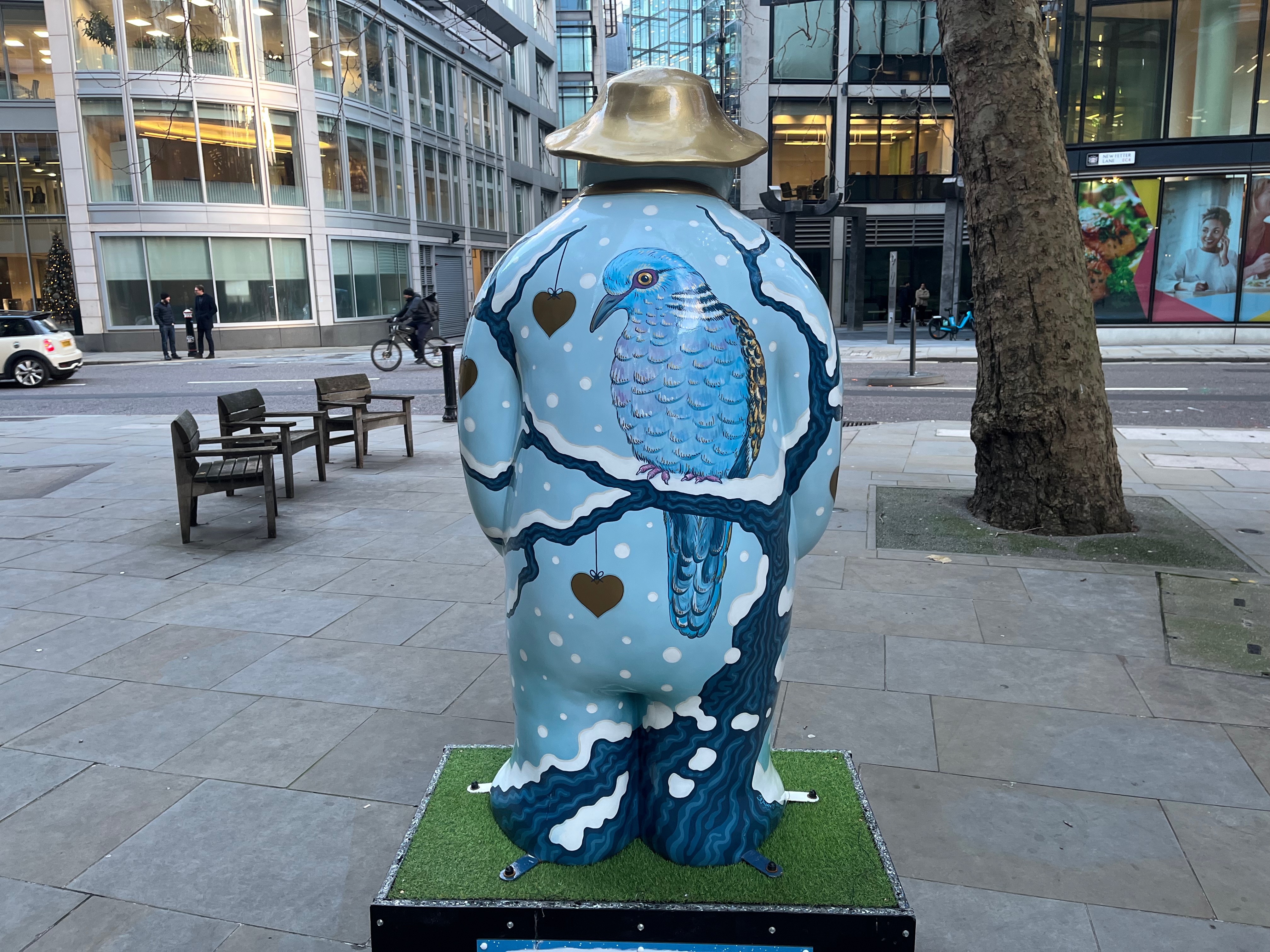 The back of The Snowman sculpture called Two Turtle Doves, showing a blue turtle dove facing us as it perches in a snow-covered tree, with a few red hearts hanging from other branches.