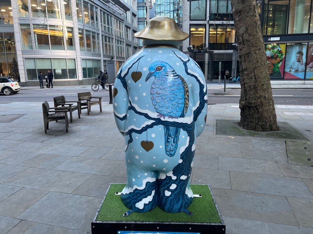 The back of The Snowman sculpture called Two Turtle Doves, showing a blue turtle dove facing us as it perches in a snow-covered tree, with a few red hearts hanging from other branches.