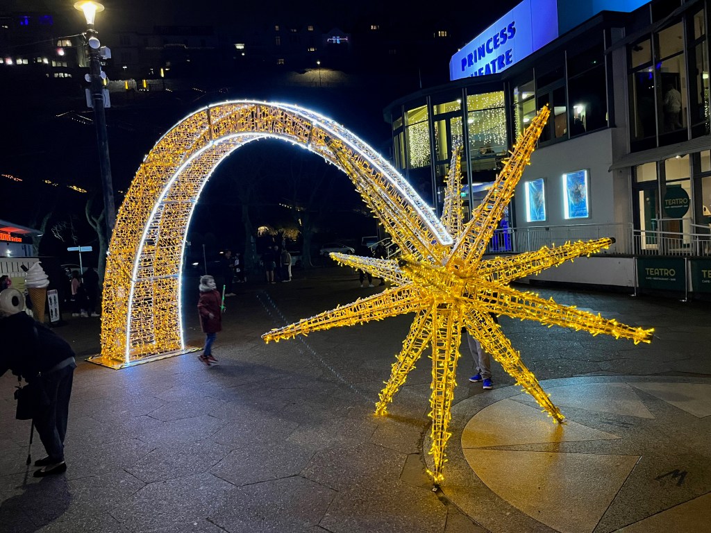 A large sculpture of a comet with a big star at one end, covered in festive lights. It forms a large archway that people can walk through, or stand under to have photos taken.