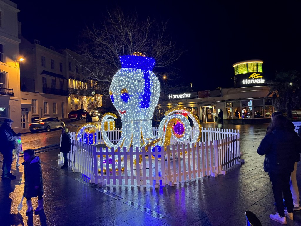 A sculpture of an octopus sitting up and smiling. Its body is covered in white festive lights, while it has round blue eyes and a small blue hat on its head.