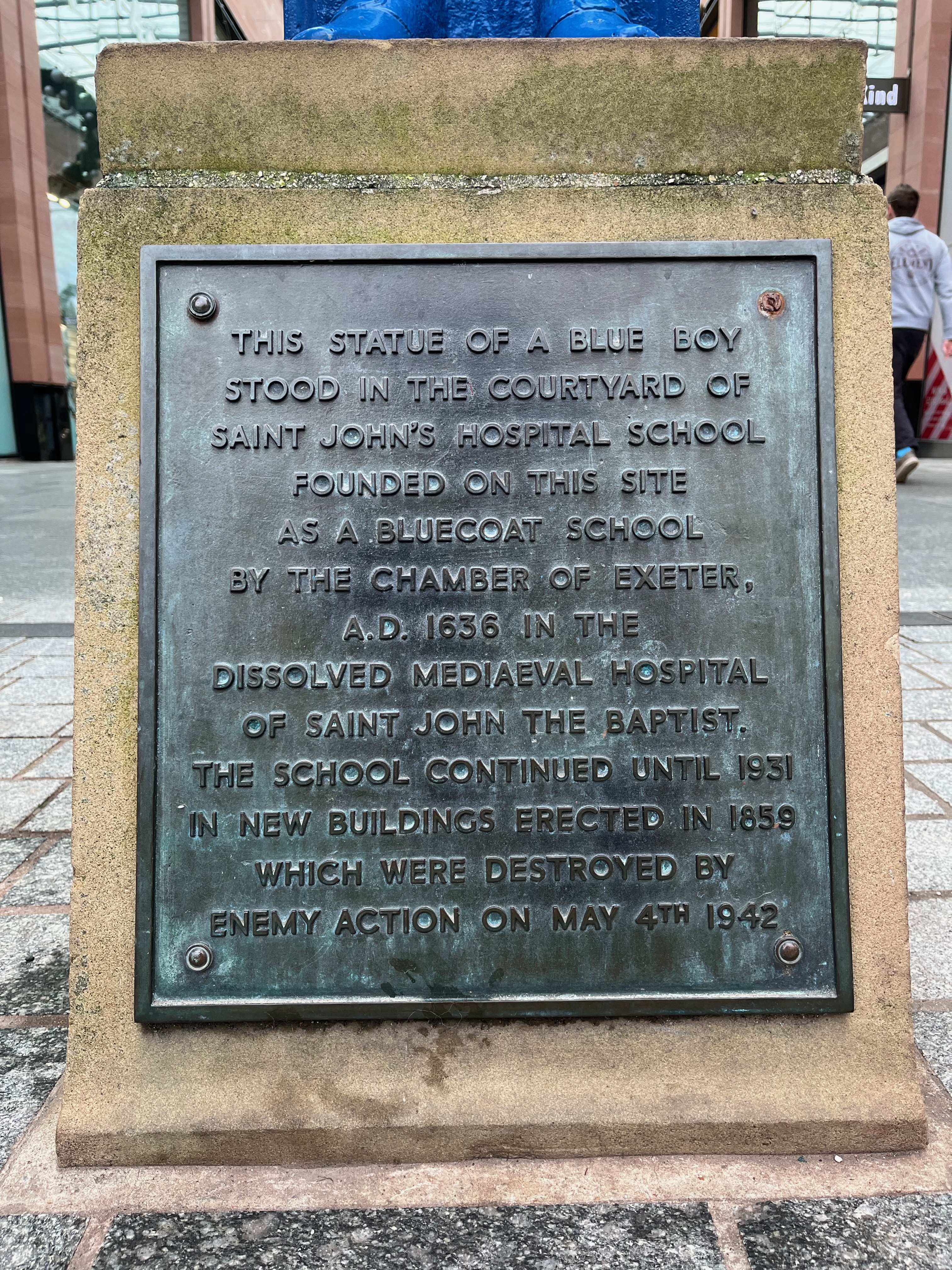 The plaque on the Blue Boy statue in the Princesshay shopping centre. The inscription reads as follows. This statue of a blue boy stood in the courtyard of Saint John's Hospital School, founded on this site as a Bluecoat school by the Chamber of Exeter, A.D. 1636 in the dissolved mediaeval hospital of Saint John the Baptist. The school continued until 1931 in the new buildings erected in 1859, which were destroyed by enemy action on May 4th 1942.