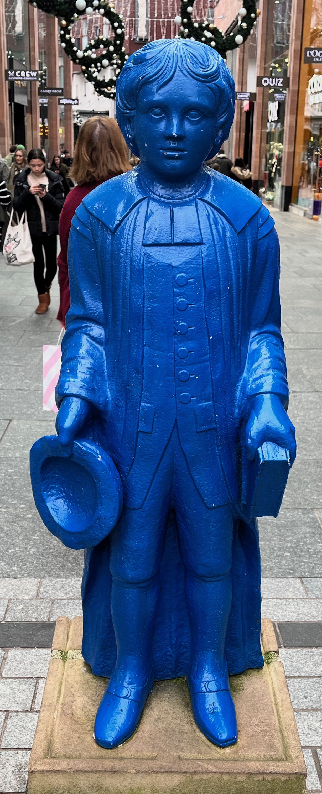 A statue of a smartly dressed young boy with wavy hair, holding a hat in one hand and book in the other, located in the Princesshay shopping centre in Exeter. The entire sculpture is a striking blue colour.