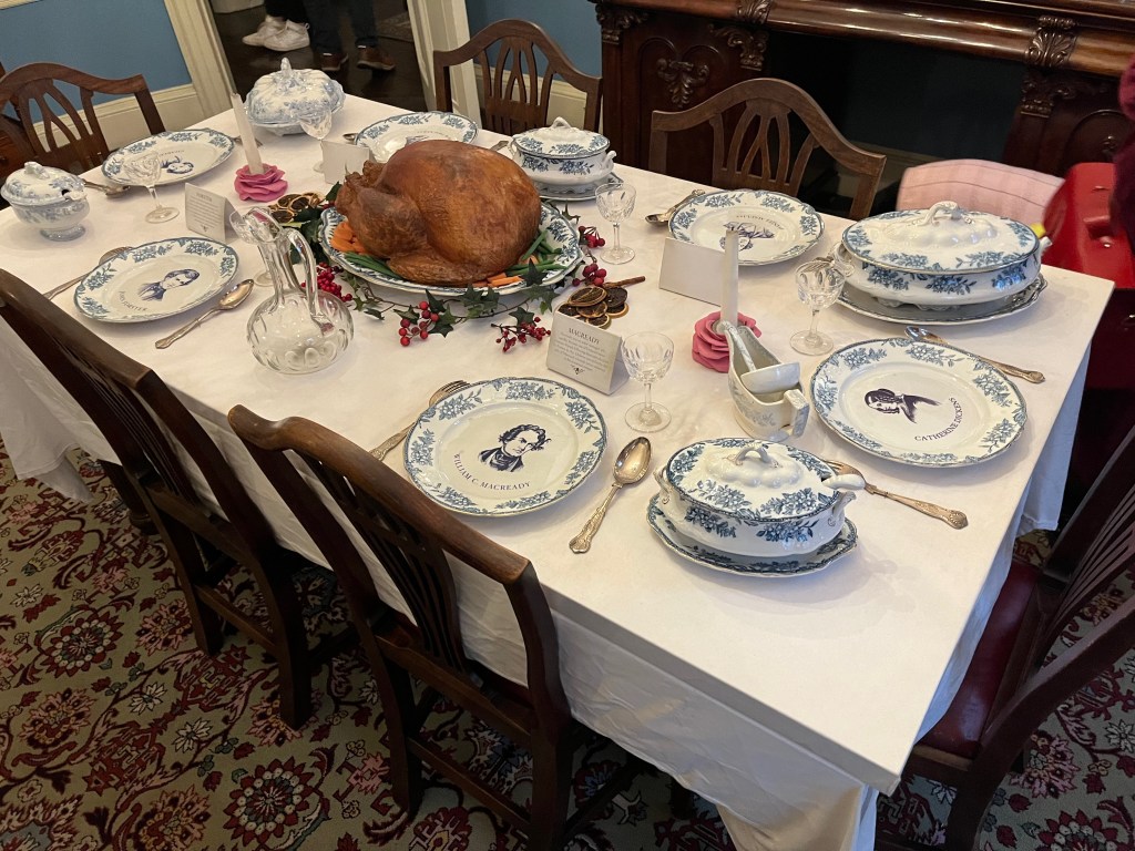 A table laid out for Christmas dinner in the Charles Dickens Museum. The white crockery has ornate blue decorations including portrait sketches of people in the centre of the plates. A model of a large cooked turkey sits on a dish in the centre of the table.